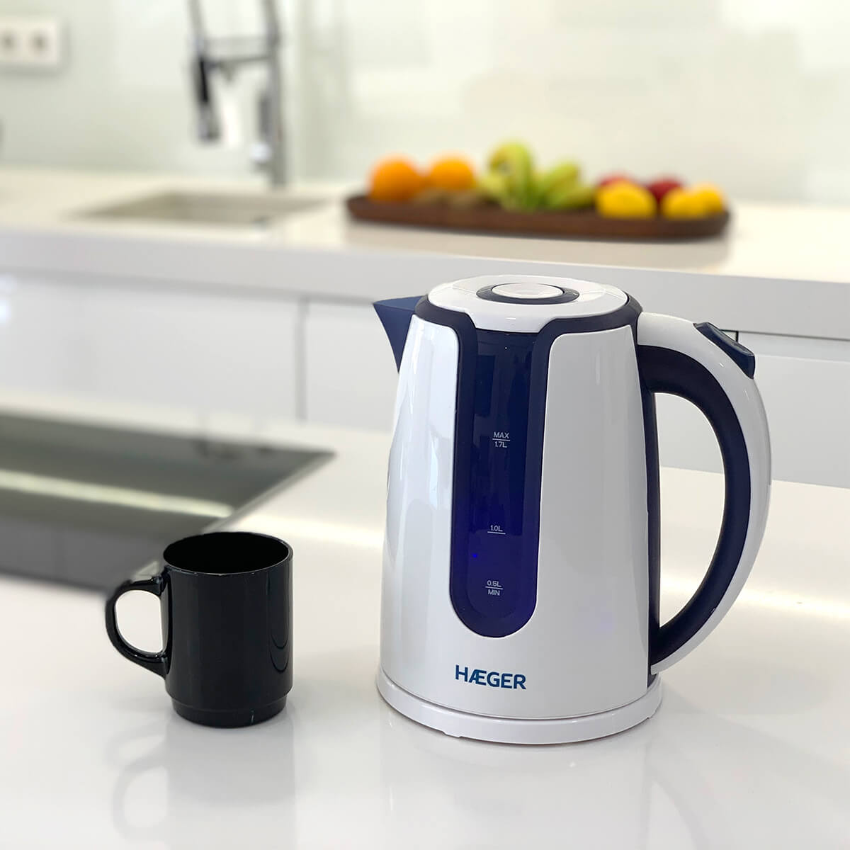 Una tetera eléctrica blanca y azul junto a una taza negra, sobre una encimera blanca.
