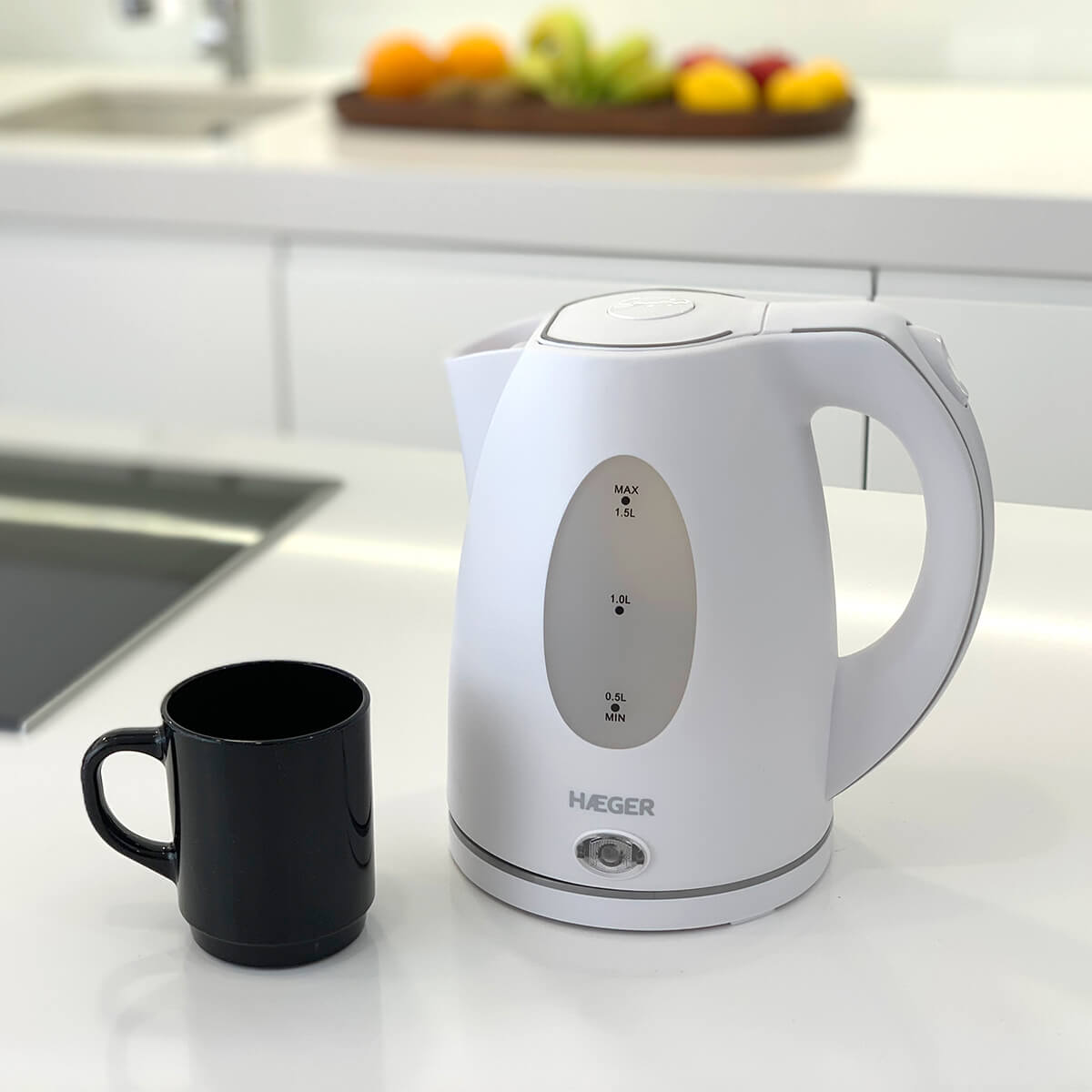 Bollitore elettrico bianco e una tazza nera su un piano di lavoro bianco. Sfondo sfocato con cibo visibile.