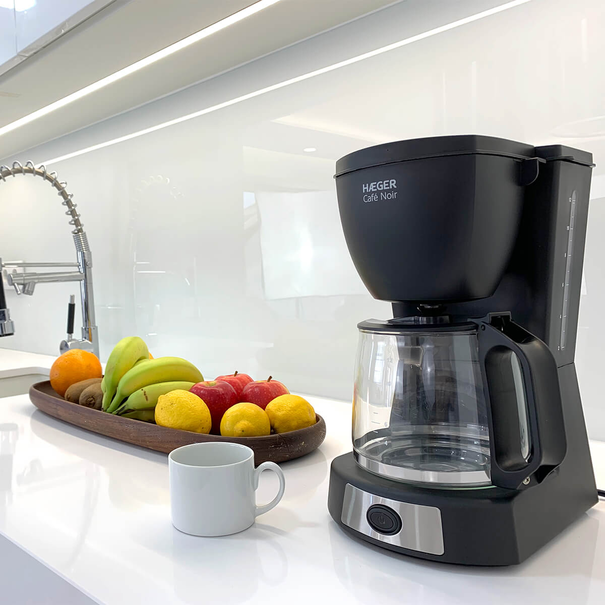 Czarny ekspres do kawy obok drewnianej tacy z owocami, z białym kubkiem na białym blacie.