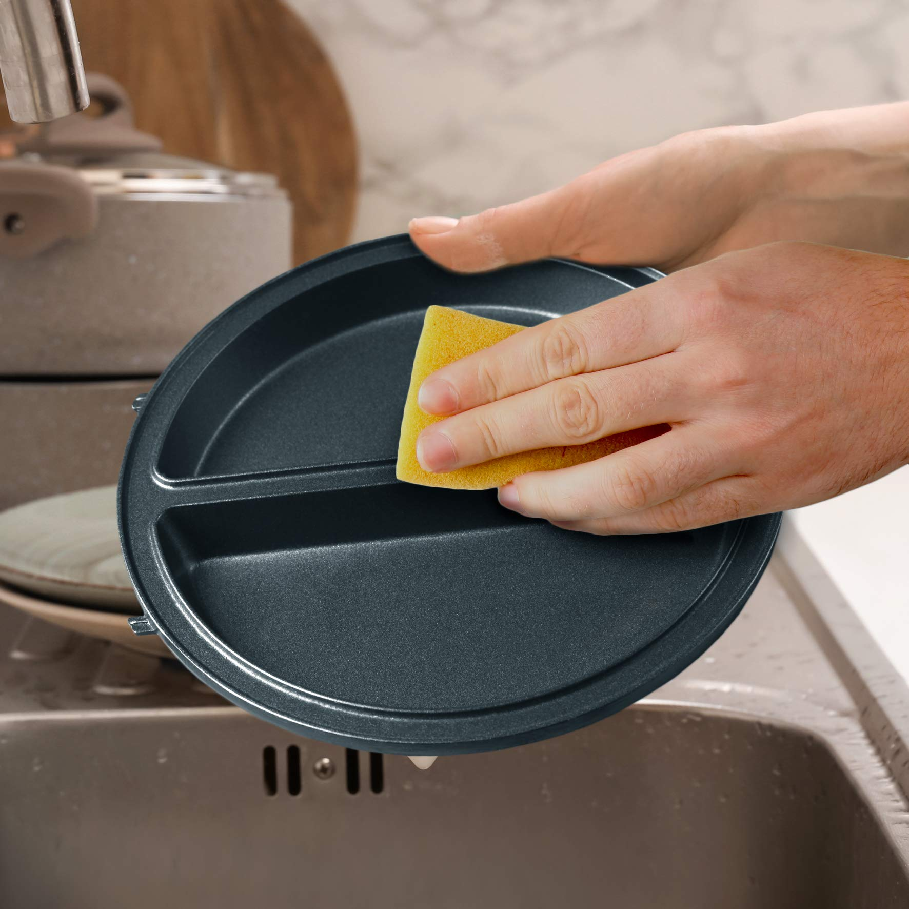 Hände waschen einen dunkelgrauen, geteilten Teller mit einem Schwamm in einem Küchenbecken.