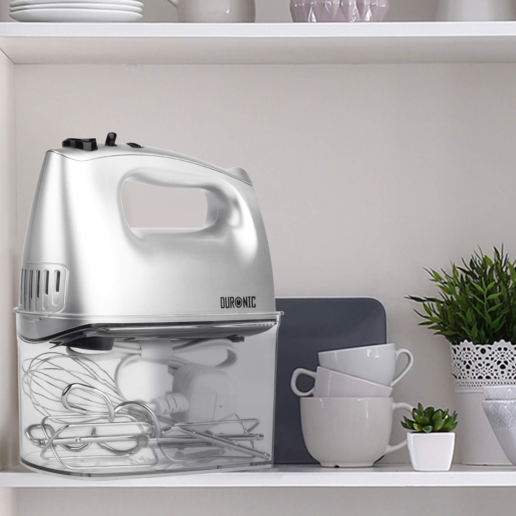 Ein weißer und silberner Handmixer mit Zubehör in einem transparenten Behälter, auf einem Regal mit Deko platziert.