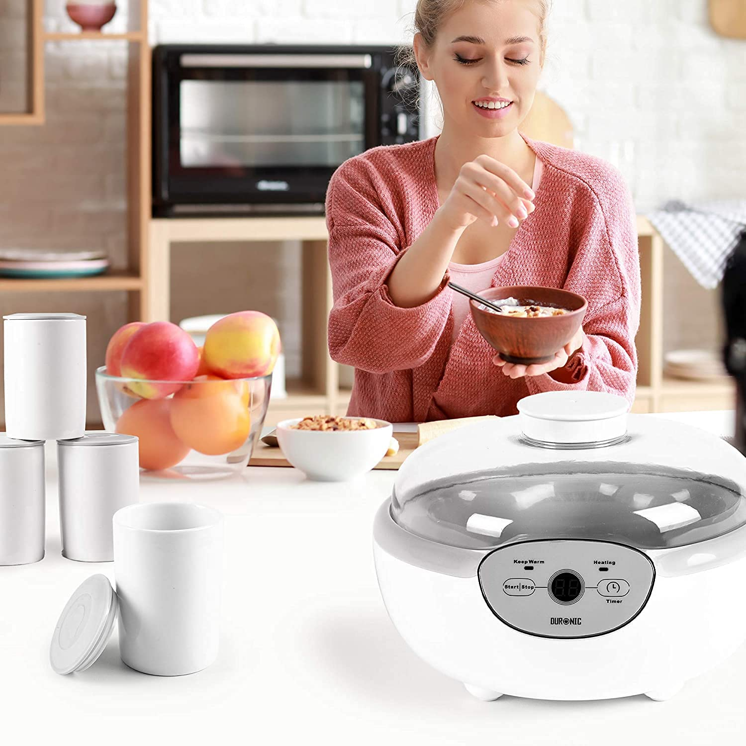 Mujer preparando una comida con un calienta alimentos y tazas blancas. Frutas en la mesa.