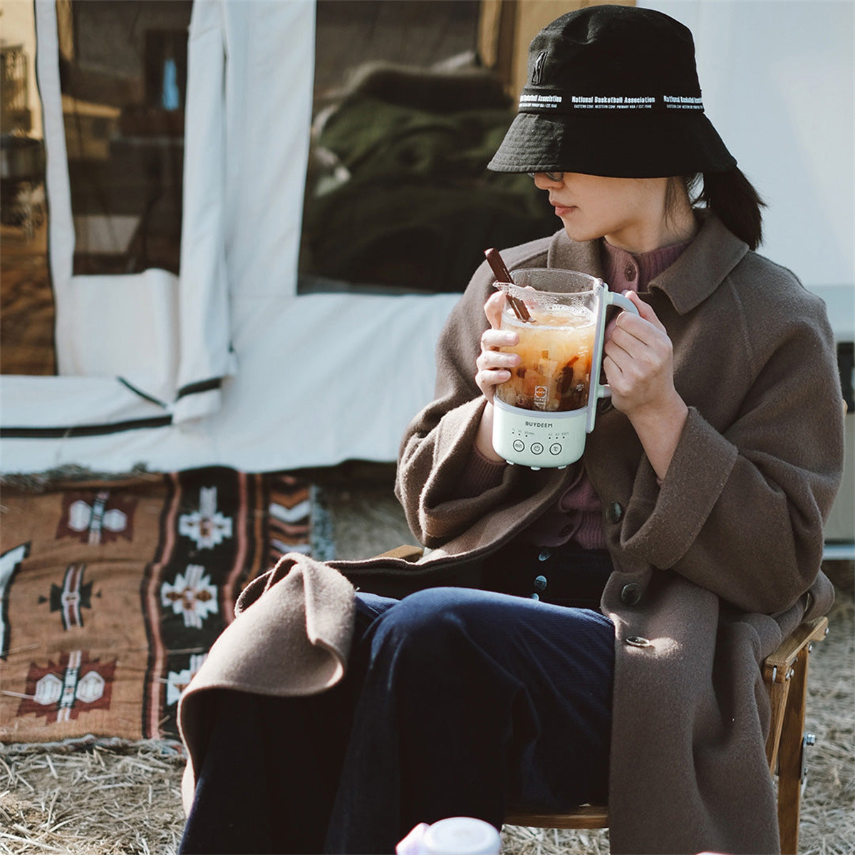 Osoba w kapeluszu i płaszczu trzymająca szklankę z napojem i małe białe urządzenie.