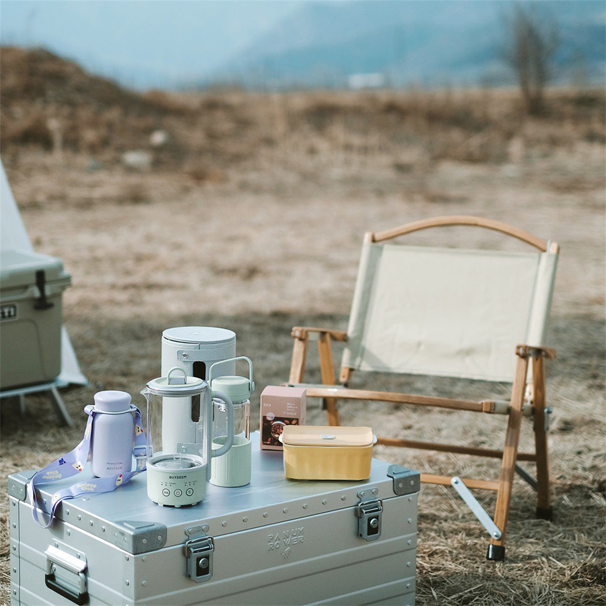 Stolik kempingowy z różnymi przedmiotami outdoorowymi i składanym krzesłem na trawiastym polu, z górami.