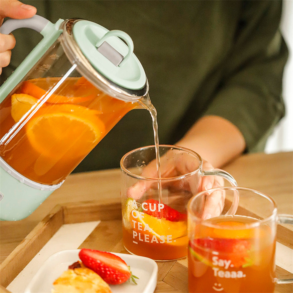 Osoba nalewa herbatę pomarańczową ze szklanego czajnika do dwóch kubków z owocami i tekstem.