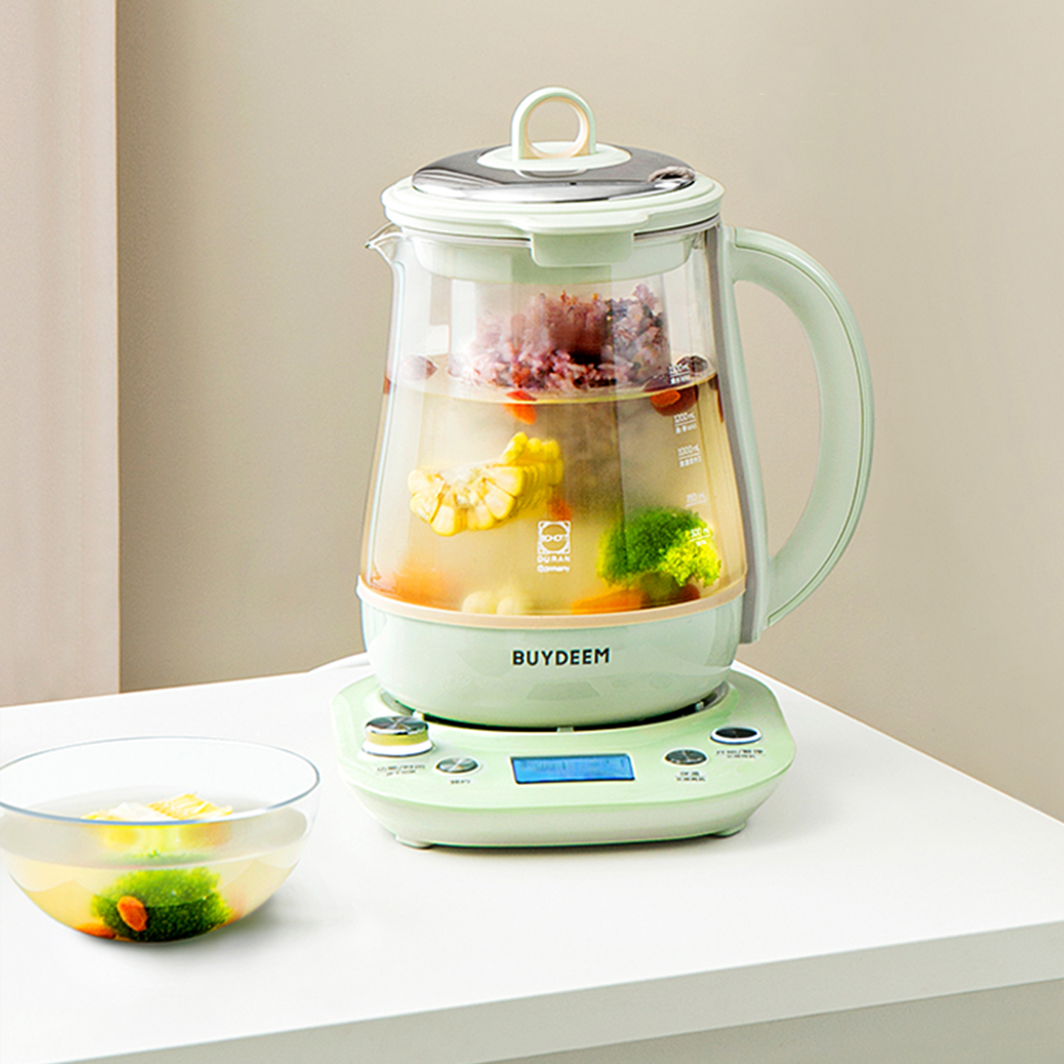 Szklany czajnik na białej powierzchni, wypełniony warzywami i wodą. Zielona podstawa z panelem sterowania.