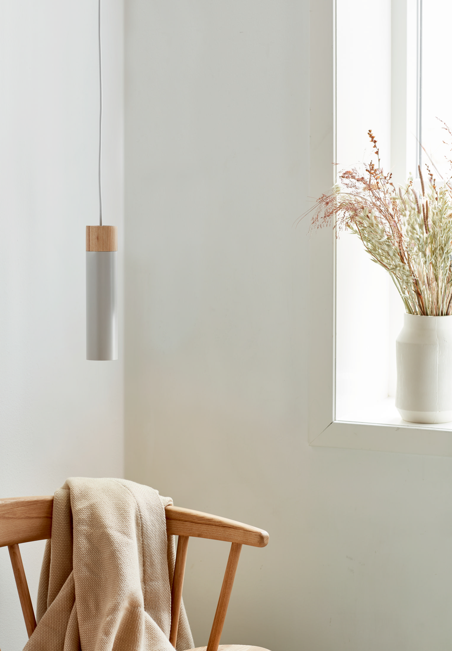 Eine Pendelleuchte hängt. Eine Vase mit getrockneten Pflanzen steht am Fenster, mit einem Holzstuhl und einer Decke im Bild.