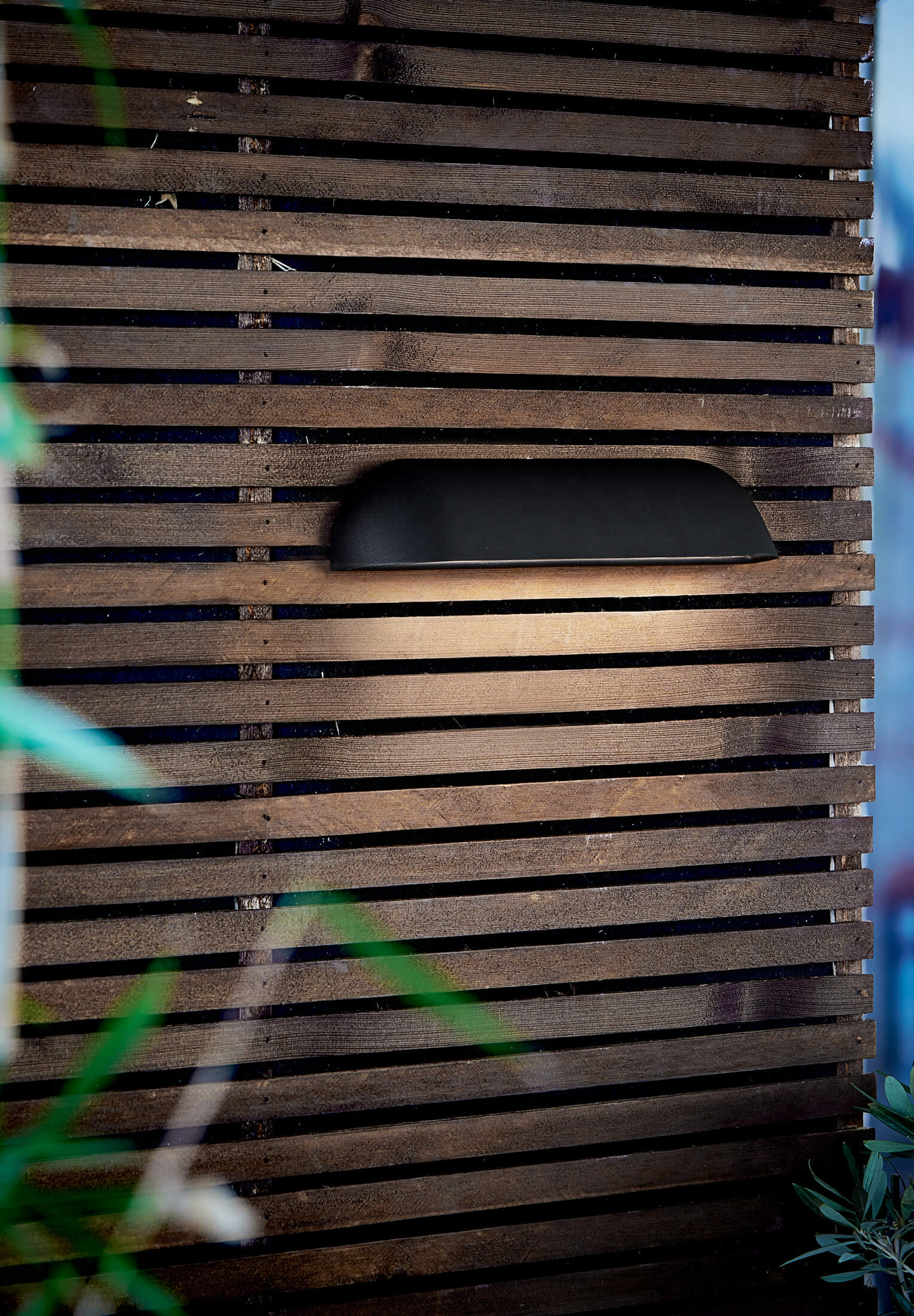 Eine schwarze Außenlampe an einer Holzlattenwand montiert. Das Licht scheint nach unten.