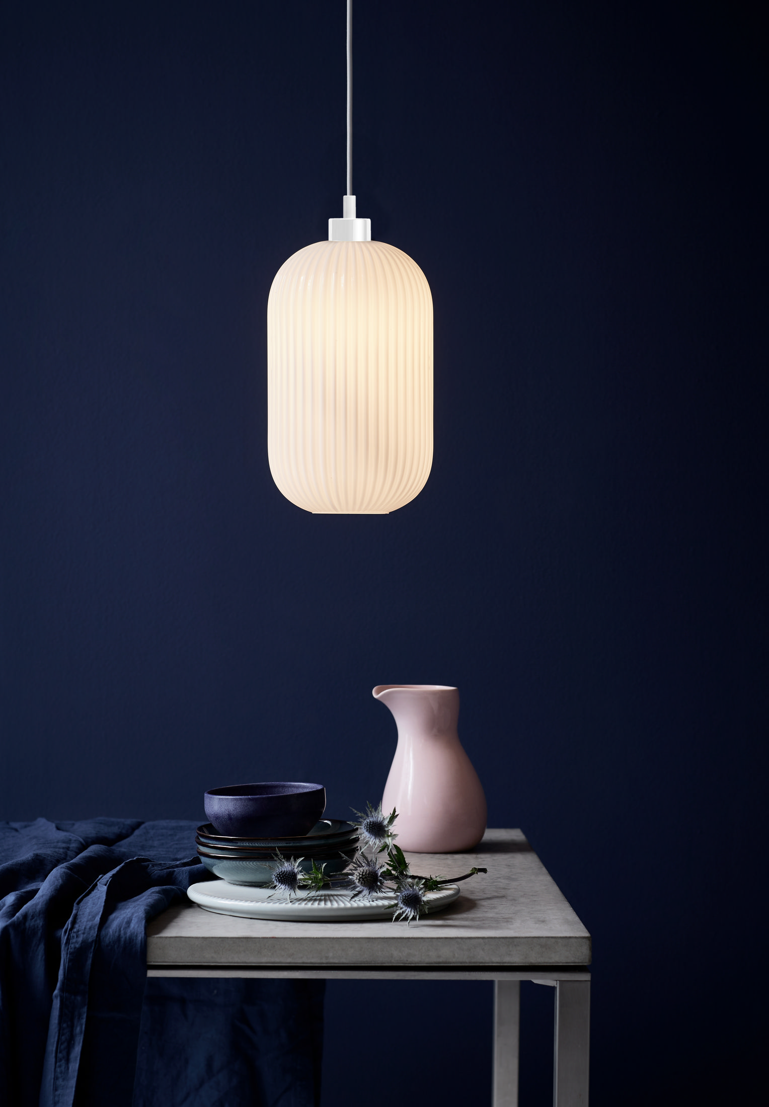 Suspension blanche avec un abat-jour ovale et nervuré au-dessus d'une table avec un vase rose et de la vaisselle bleue.