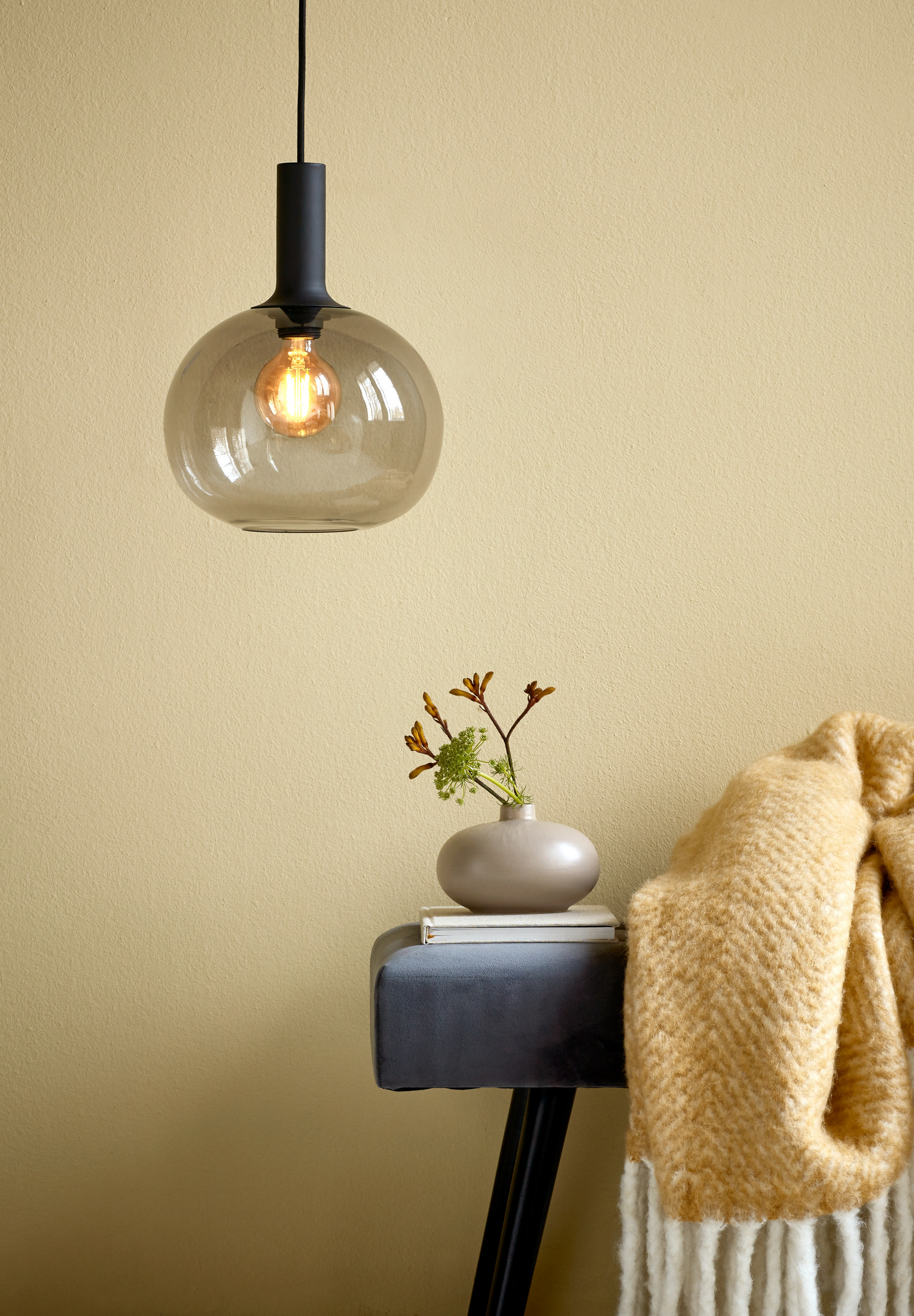 Suspension avec globe en verre au-dessus d'un vase avec verdure et couverture sur un tabouret.
