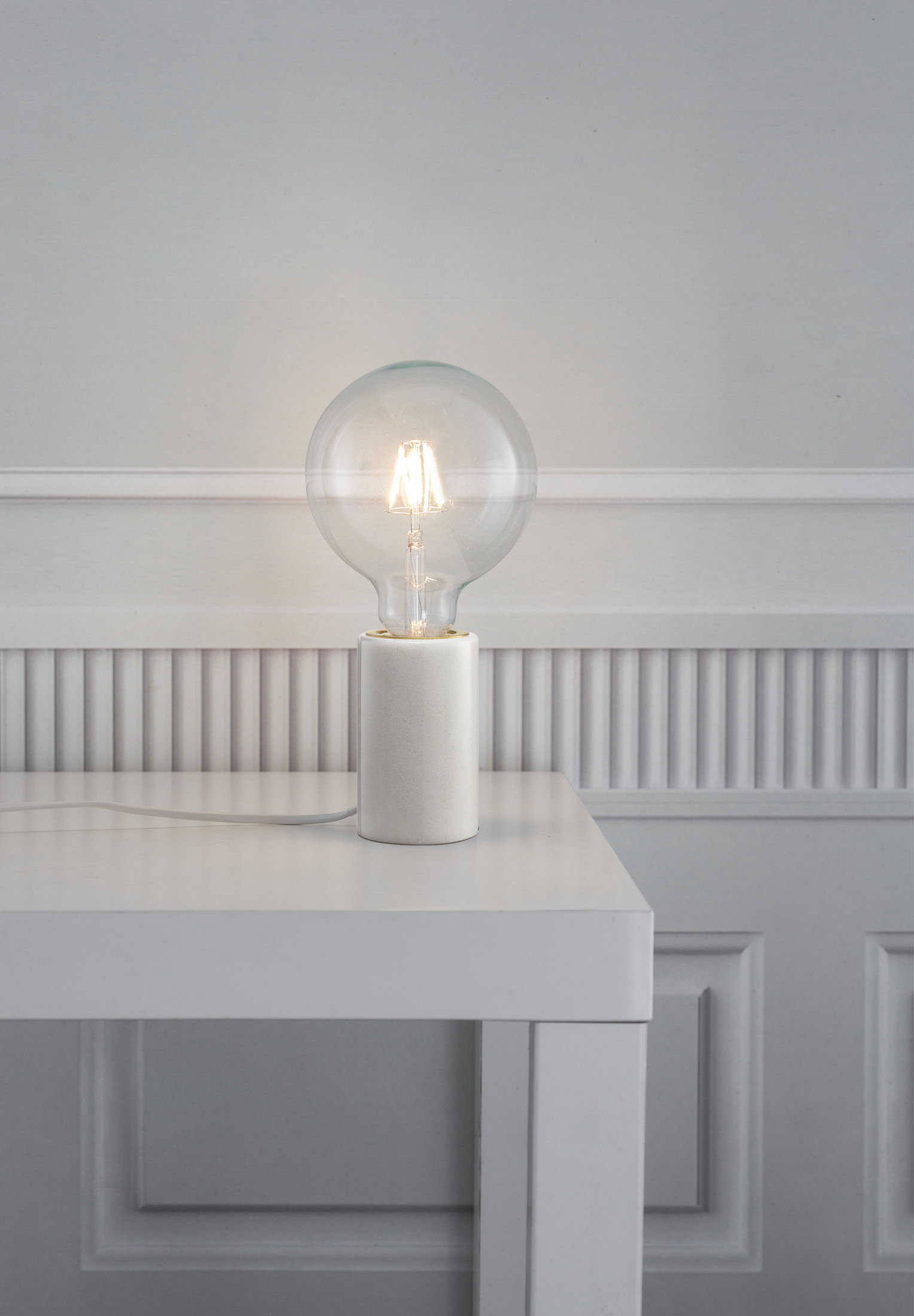 Eine weiße Tischlampe mit einer großen, klaren Glühbirne auf einem weißen Tisch, weißer Wandhintergrund.