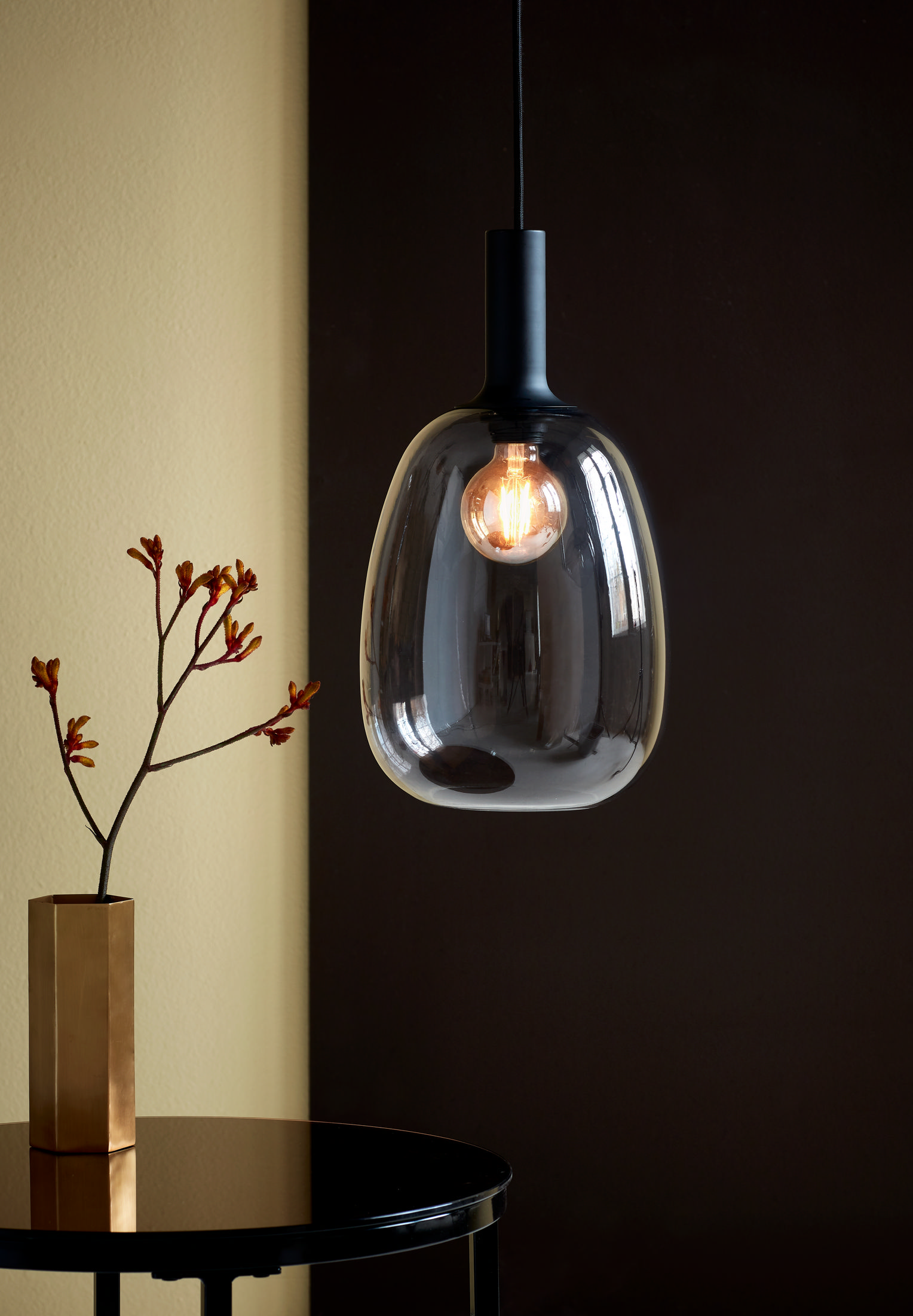 Pendelleuchte. Gläserne Tropfenform, Glühbirne innen. Dunkler Hintergrund, goldene Vase, Pflanze.