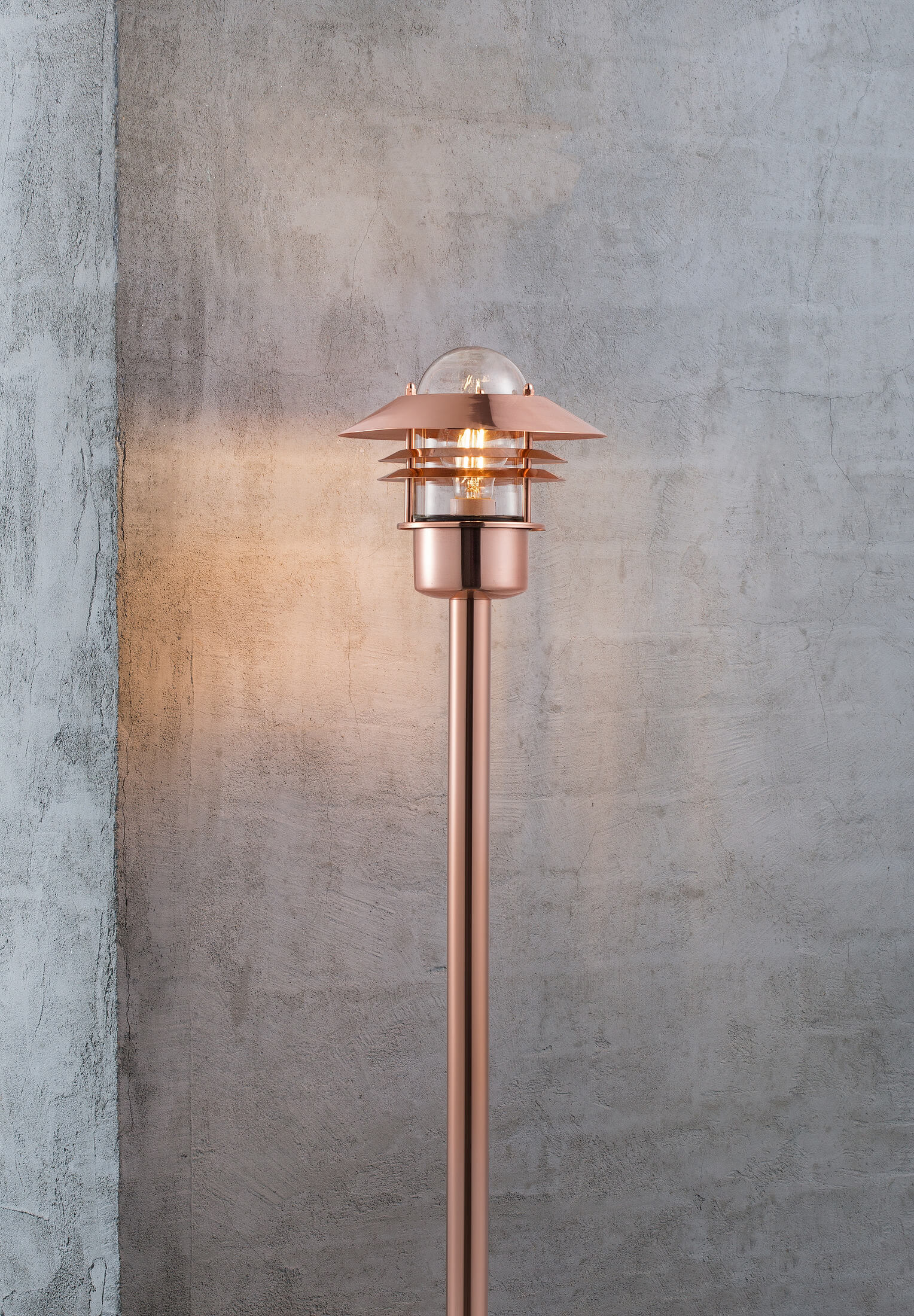 Kupferfarbene Außenlampe an einem Pfahl. Der Hintergrund ist eine graue Betonmauer.