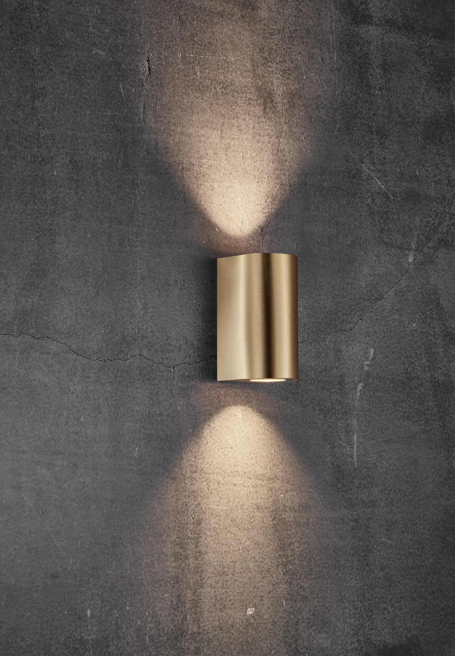 Eine zylindrische goldene Wandleuchte an einer grauen Wand. Licht scheint von der Lampe nach oben und unten.