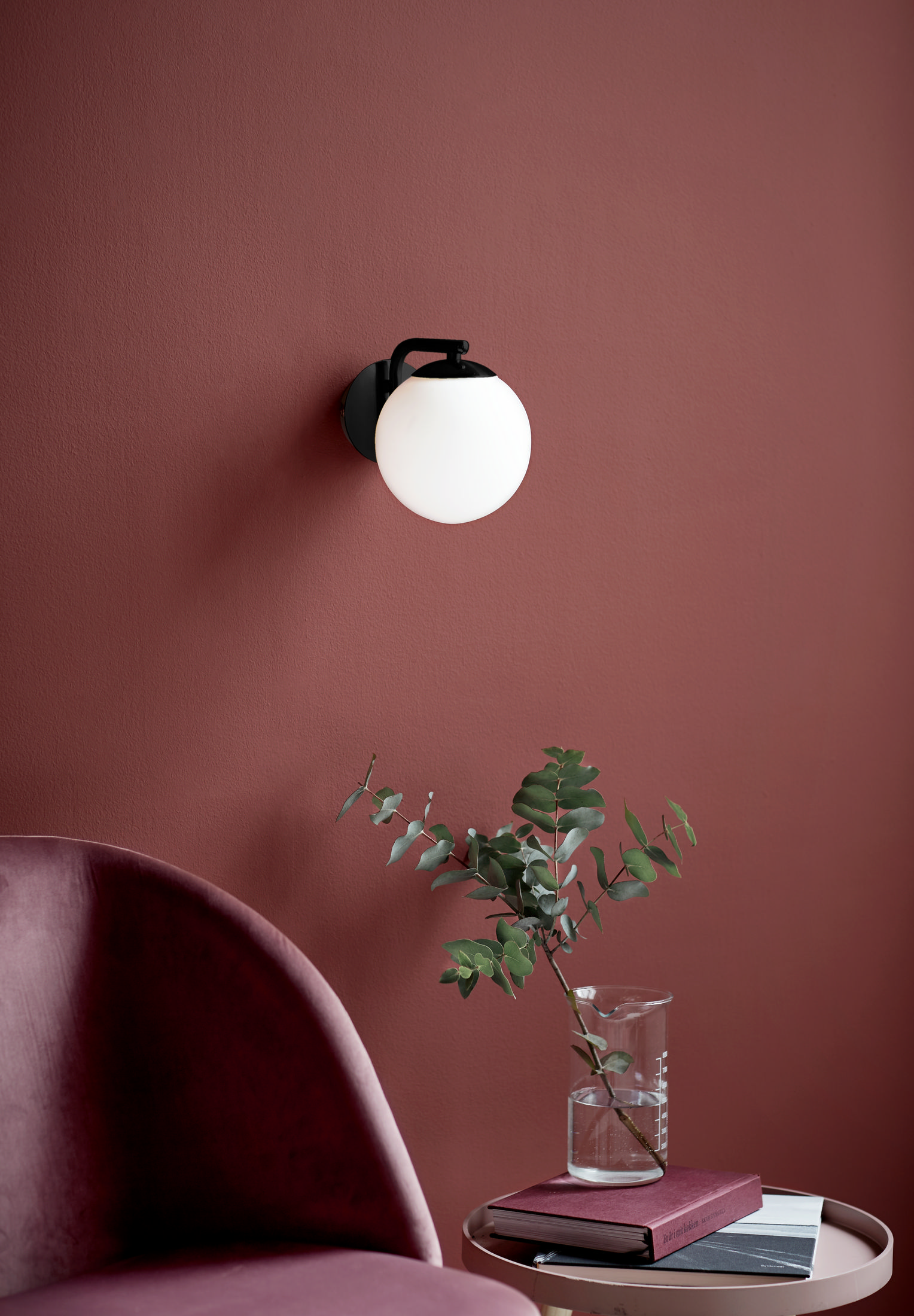 Weiße Kugellampe an einer weinroten Wand, mit einer Vase mit Grün.