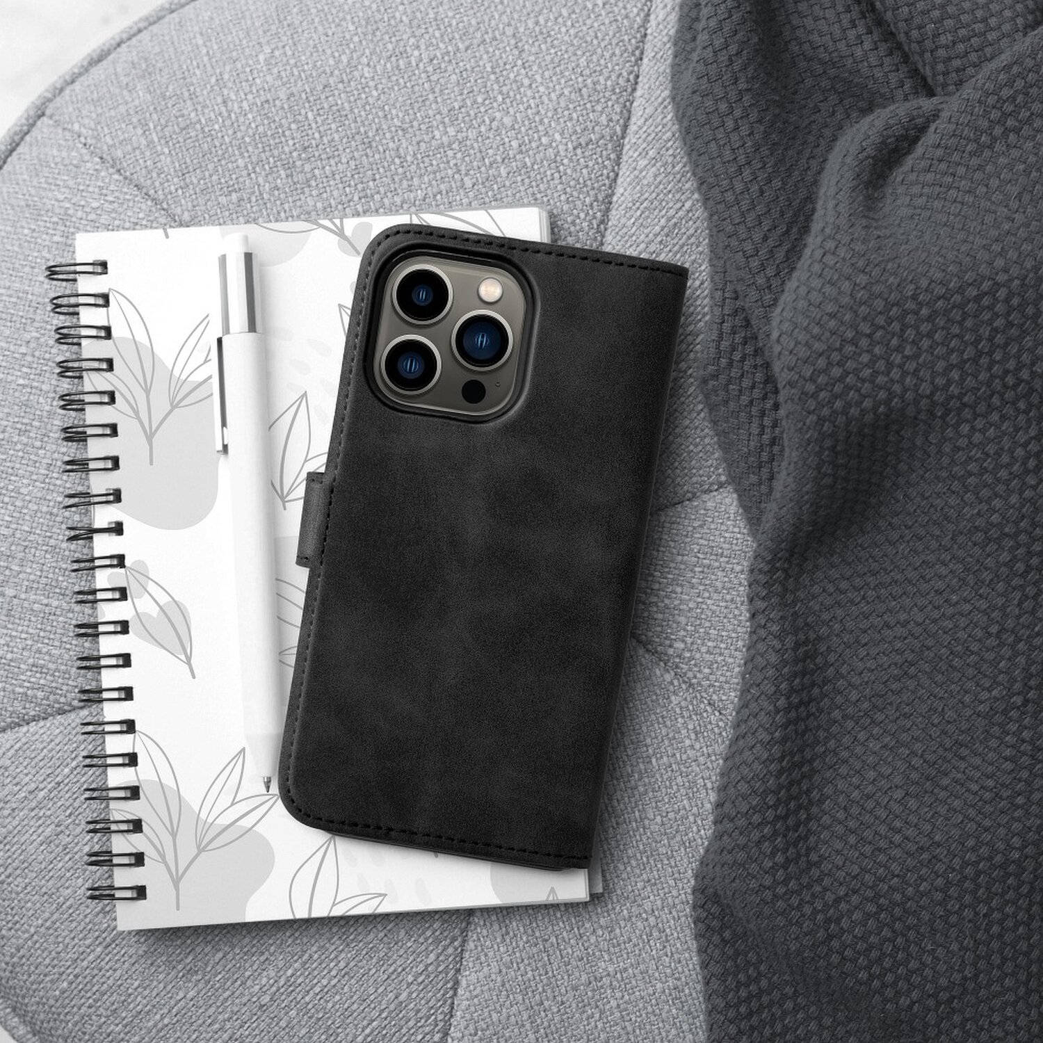 Telefono nero in una custodia nera su un taccuino e una coperta. Penna bianca sul taccuino. Sfondo grigio.
