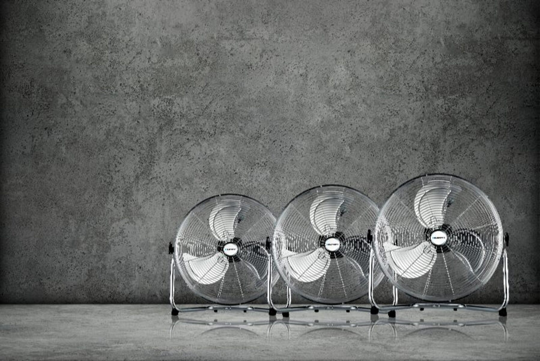 Tres ventiladores de metal en fila, contra una pared de hormigón gris.