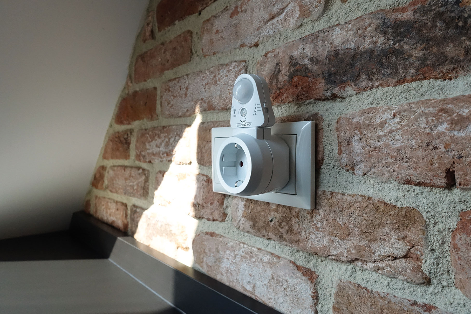 Weißer Smart-Stecker mit Sensor an einer Ziegelmauer. Schwarze Theke darunter.