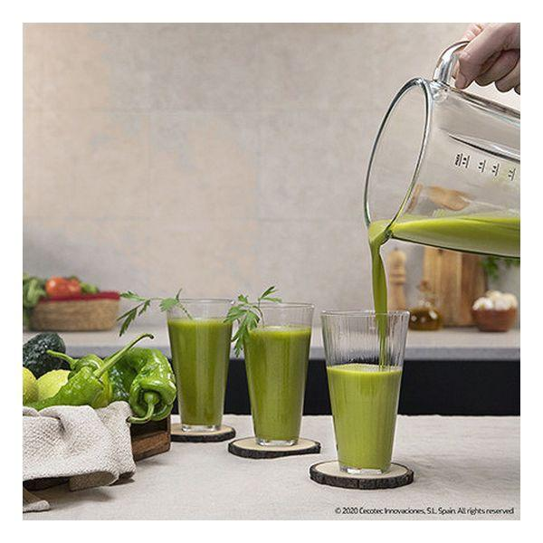 Jugo verde se vierte en vasos sobre una mesa con pimientos verdes y un posavasos de madera.