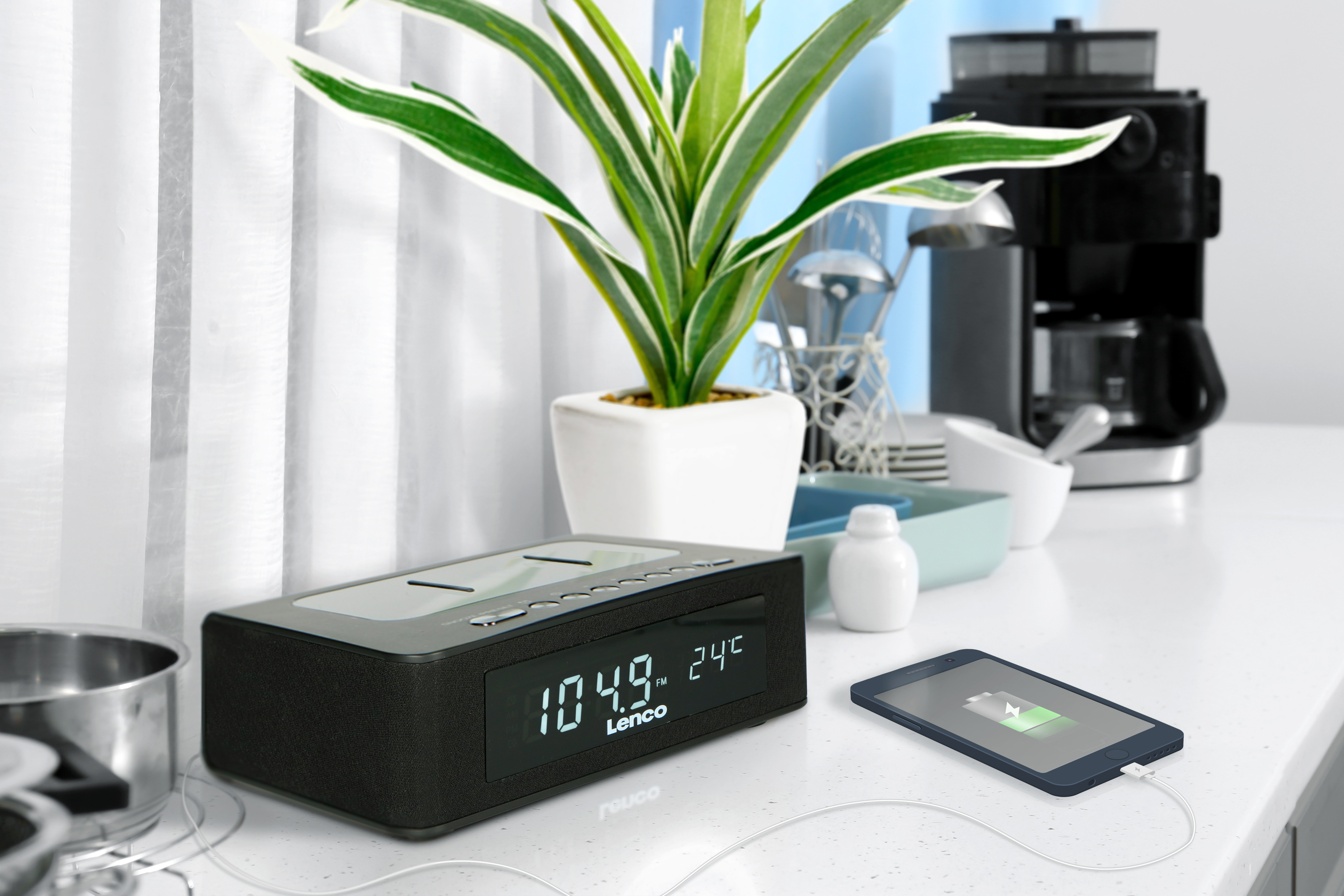 Kitchen counter with a digital clock, a plant, and a charging phone. Coffee maker in background.