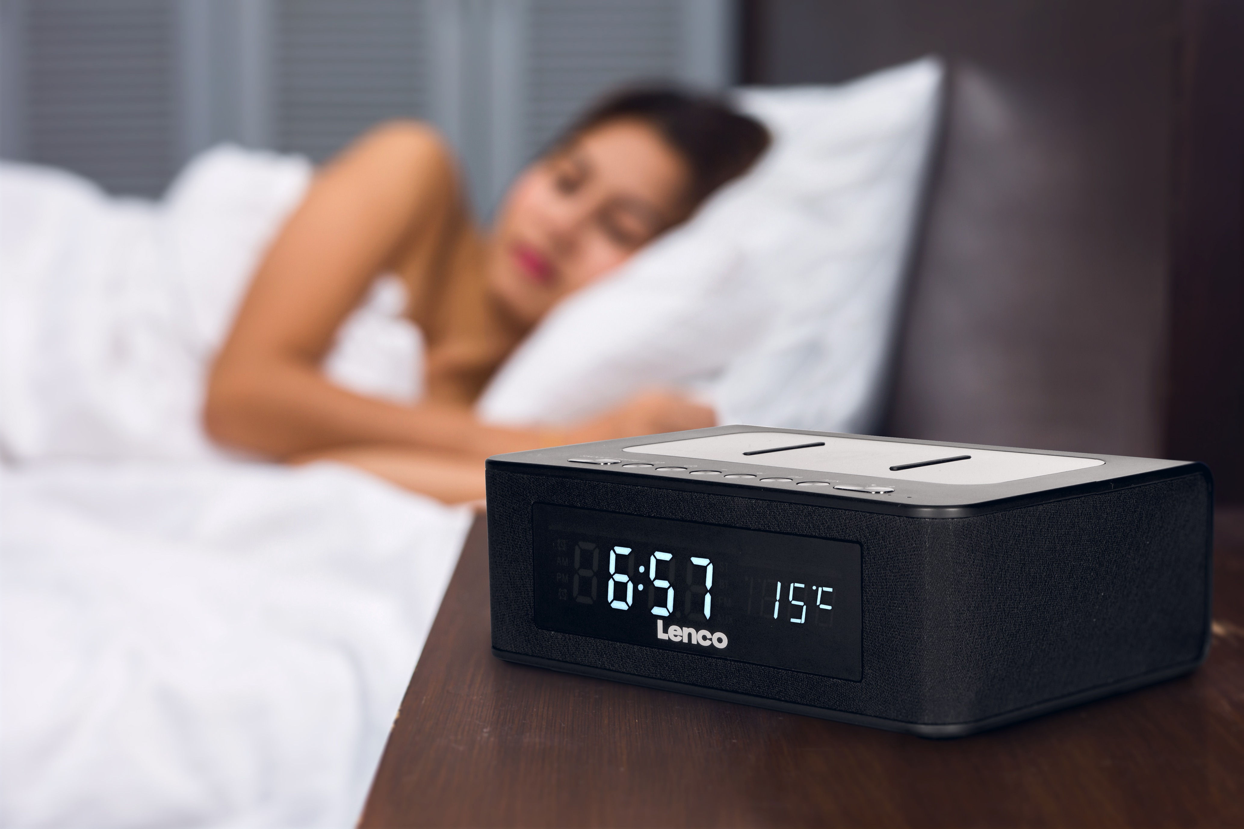 A black digital alarm clock on a nightstand next to a sleeping woman in bed.