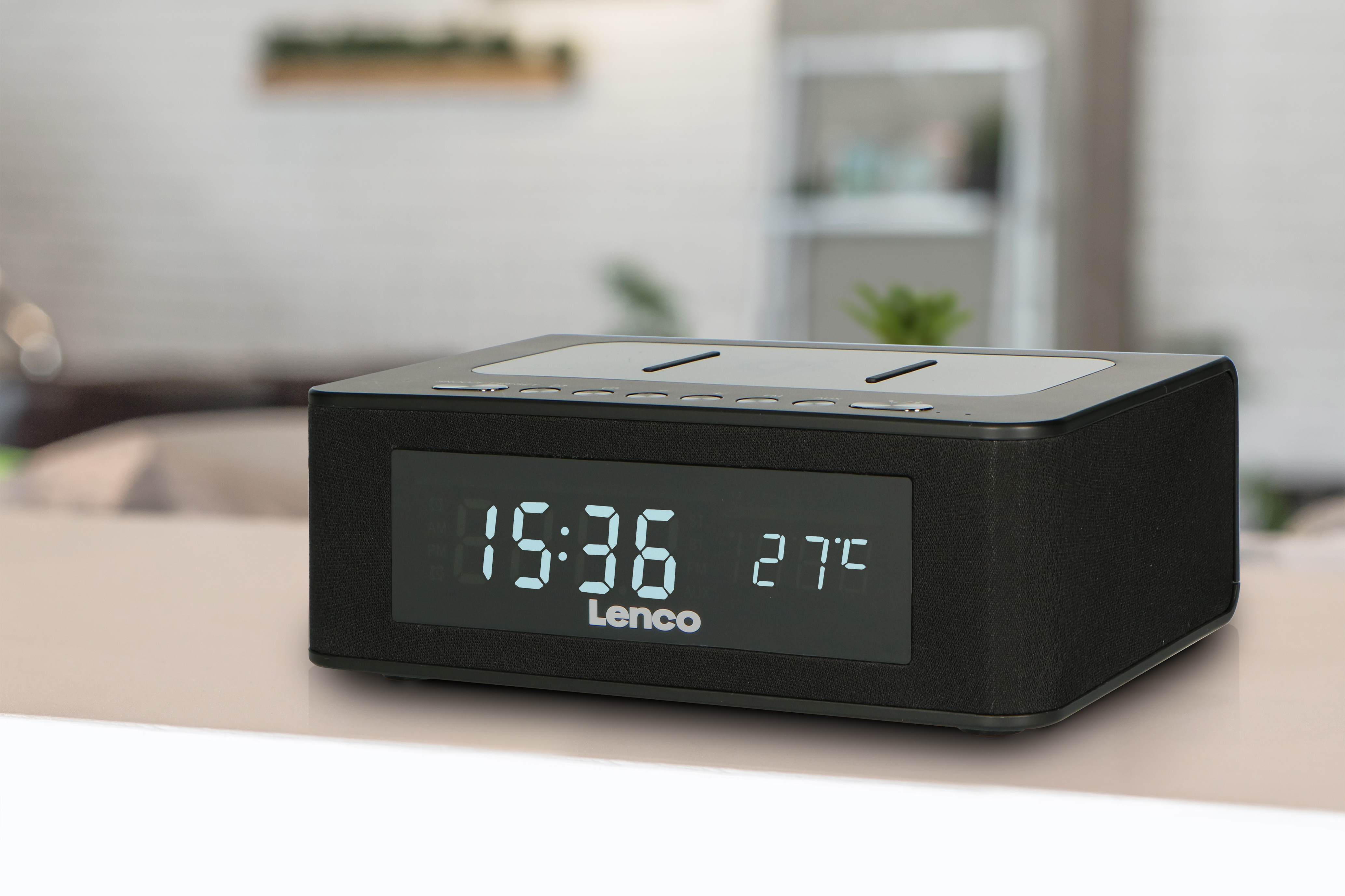 Black Lenco alarm clock displays time and temperature. It sits on a light surface, blurred background.