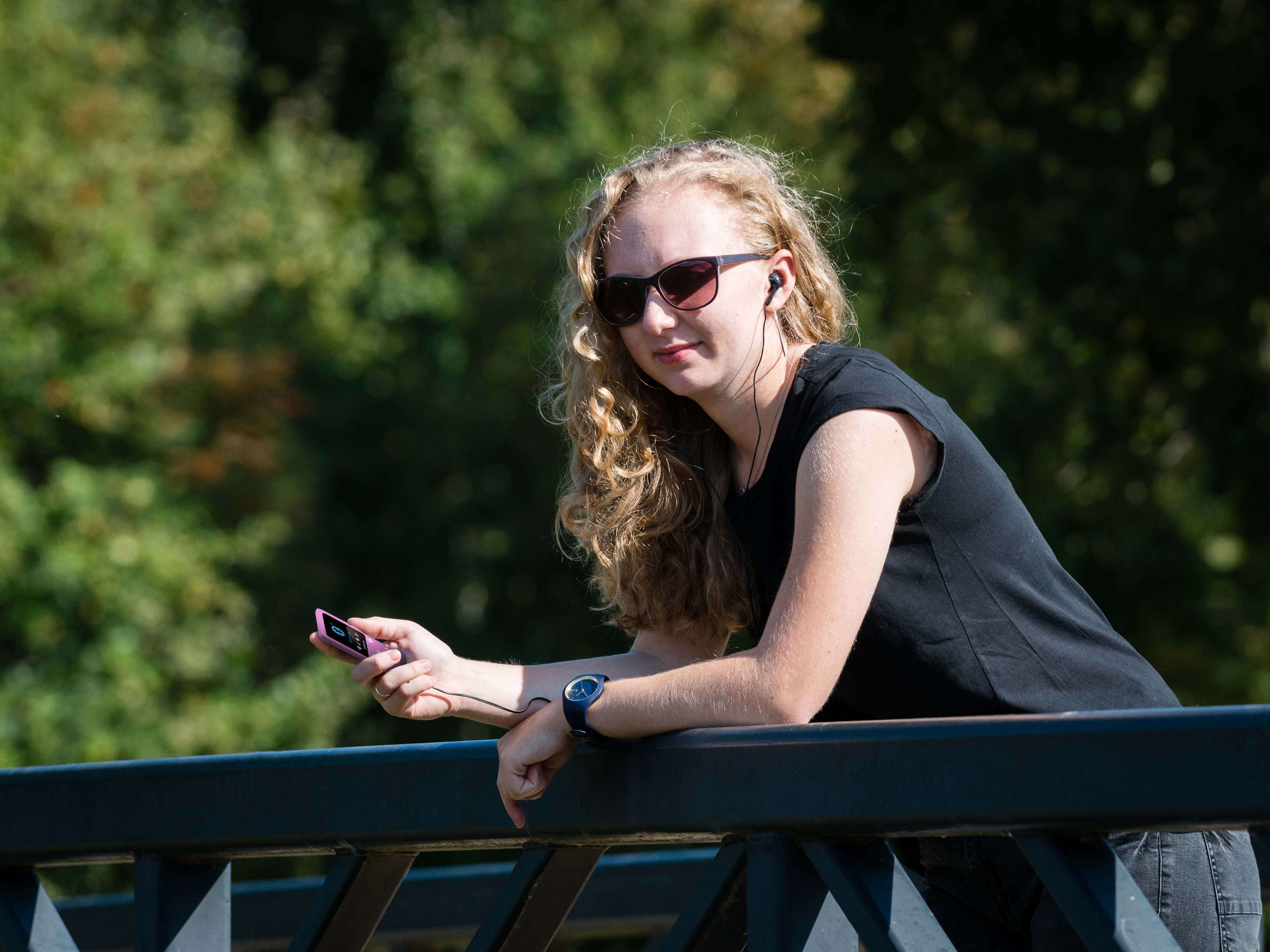 Blondynka w okularach przeciwsłonecznych i czarnym topie opiera się na balustradzie, słuchając muzyki.