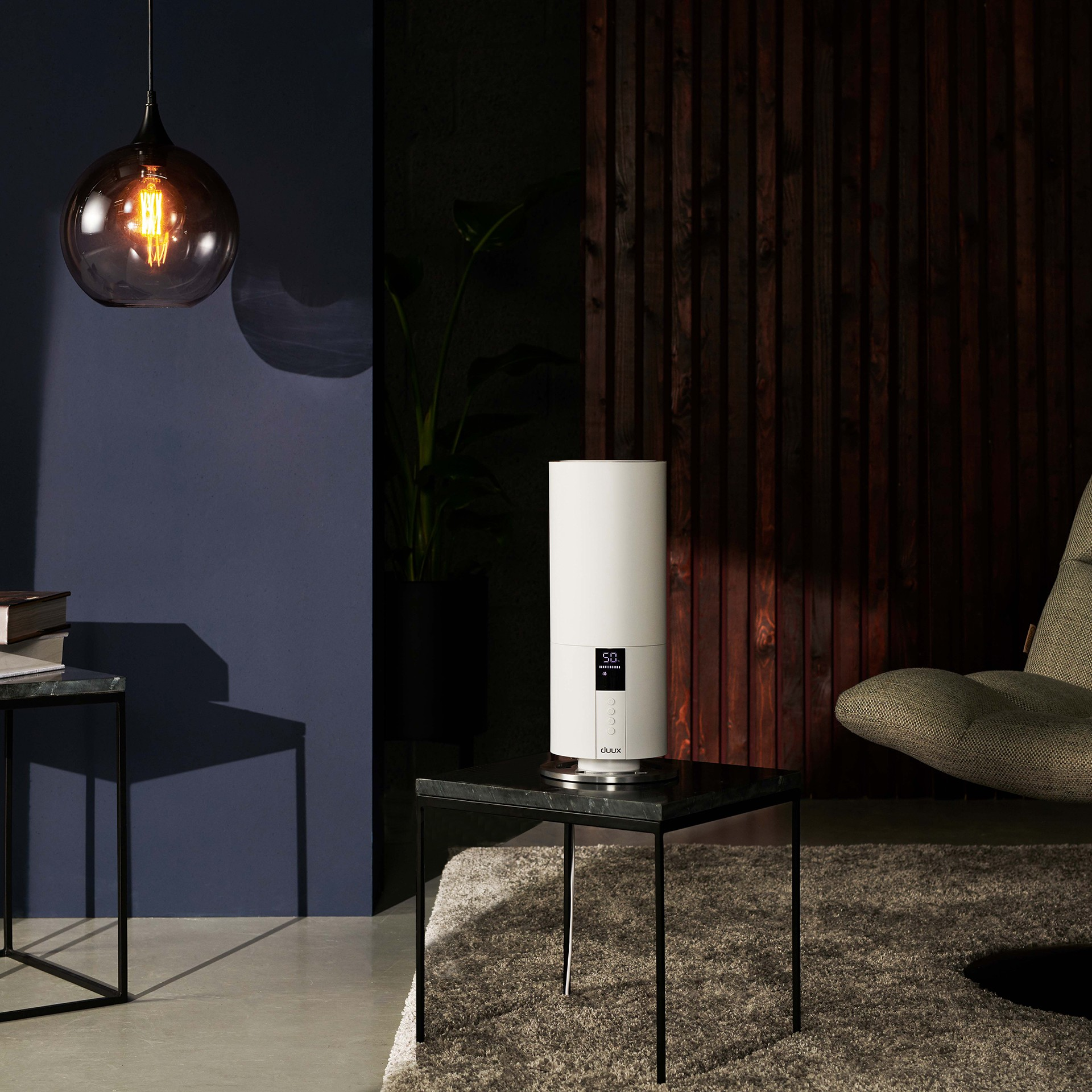 Biały nawilżacz powietrza na czarnym stoliku z lampą i rośliną w ciemnym pokoju.