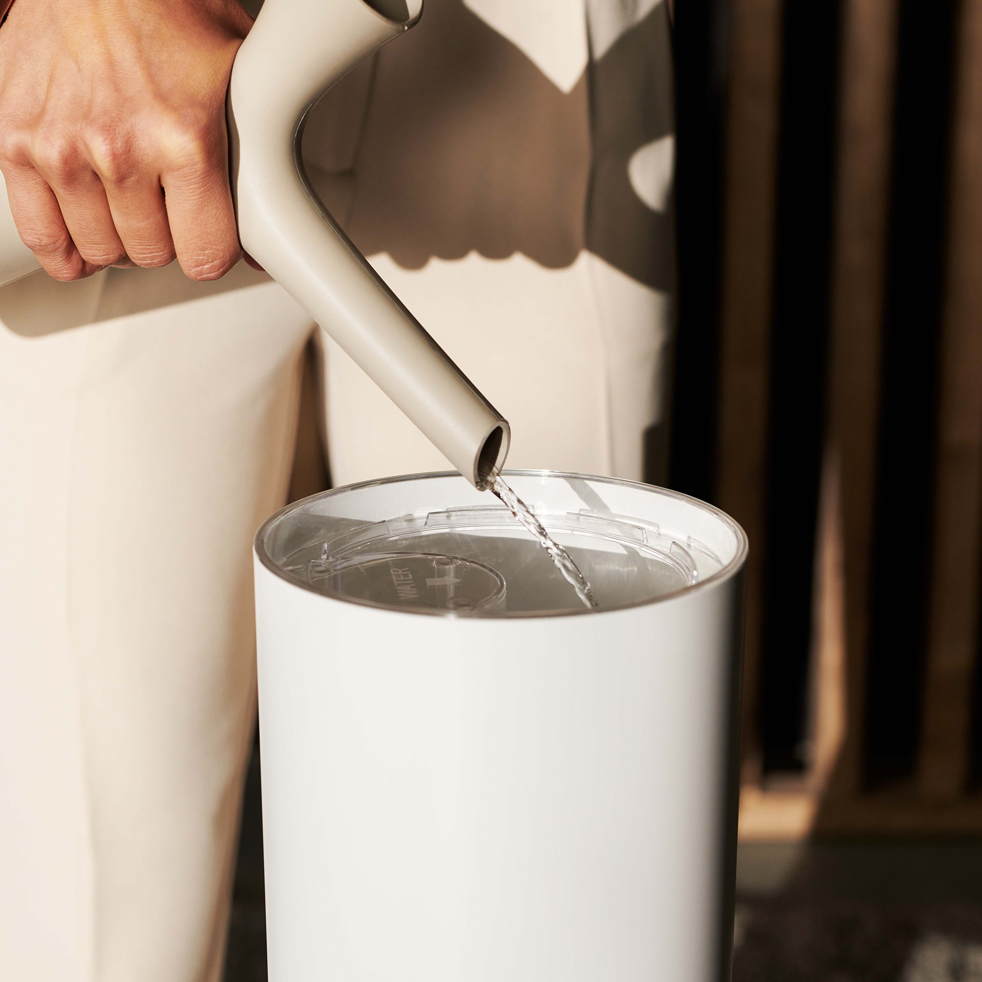 Osoba nalewa wodę z szarej konewki do białego, cylindrycznego pojemnika.