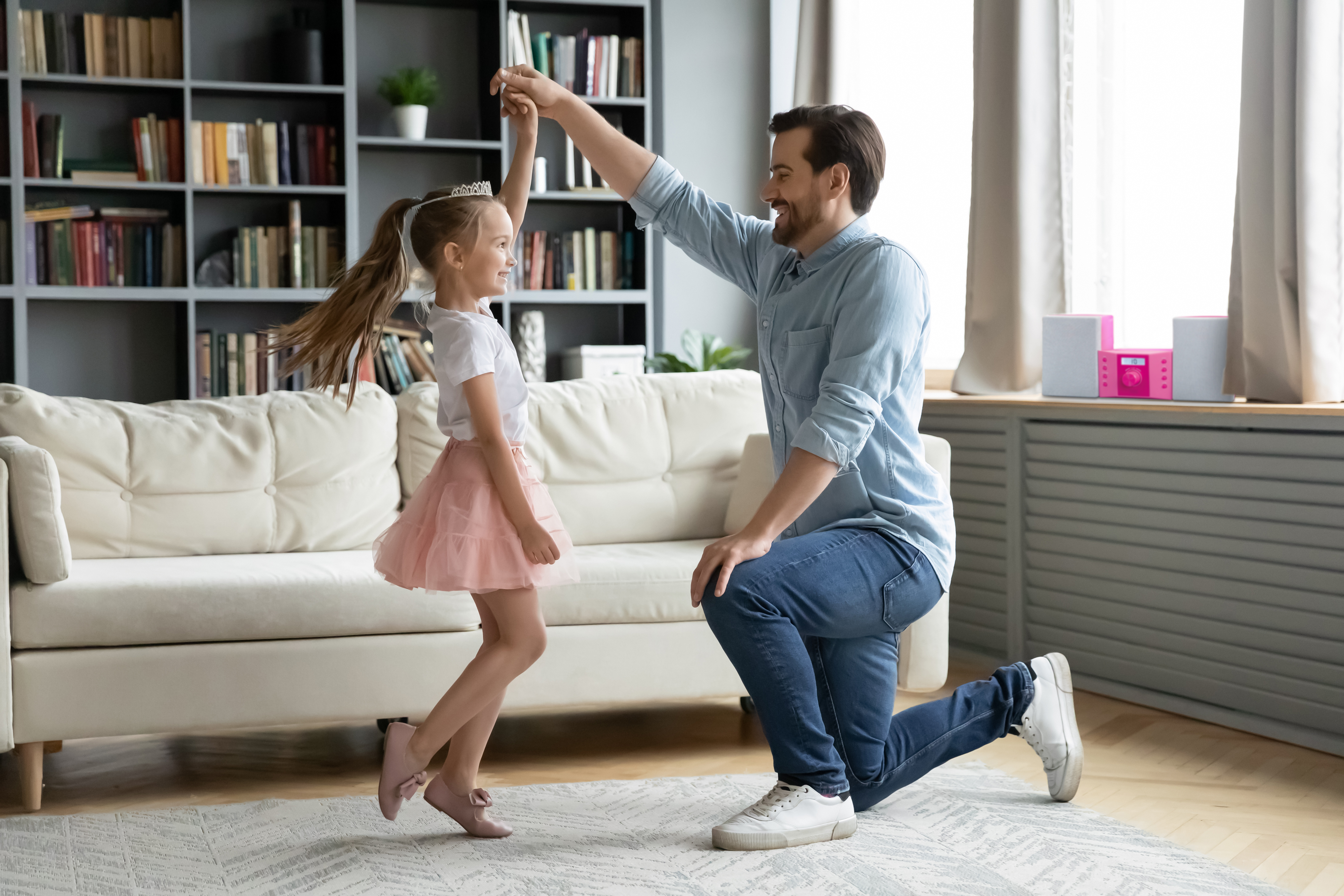 Ein Vater und eine Tochter tanzen im Wohnzimmer. Das kleine Mädchen trägt einen rosa Tutu und eine Tiara.