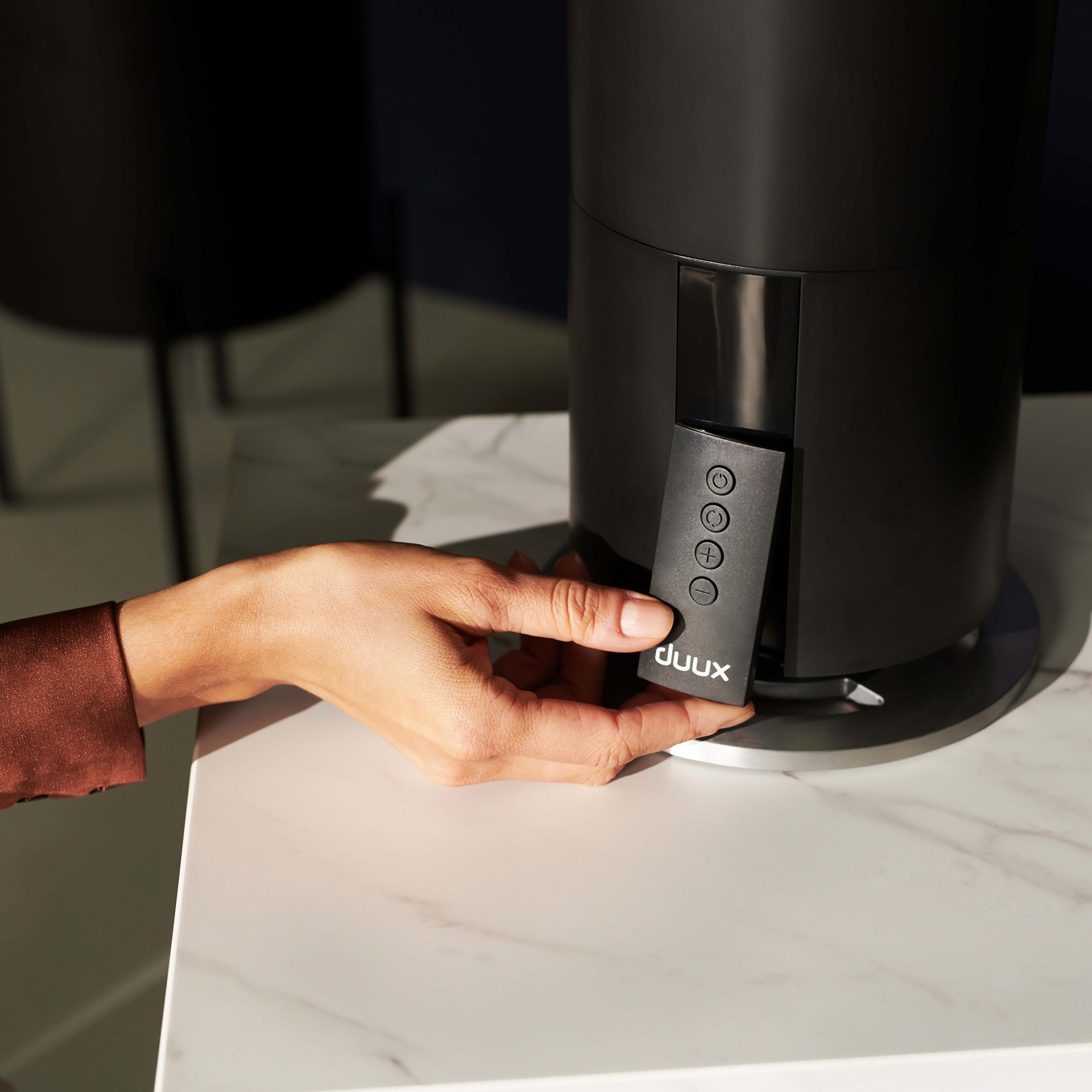 Dłoń trzymająca pilota w pobliżu czarnego cylindrycznego wentylatora.