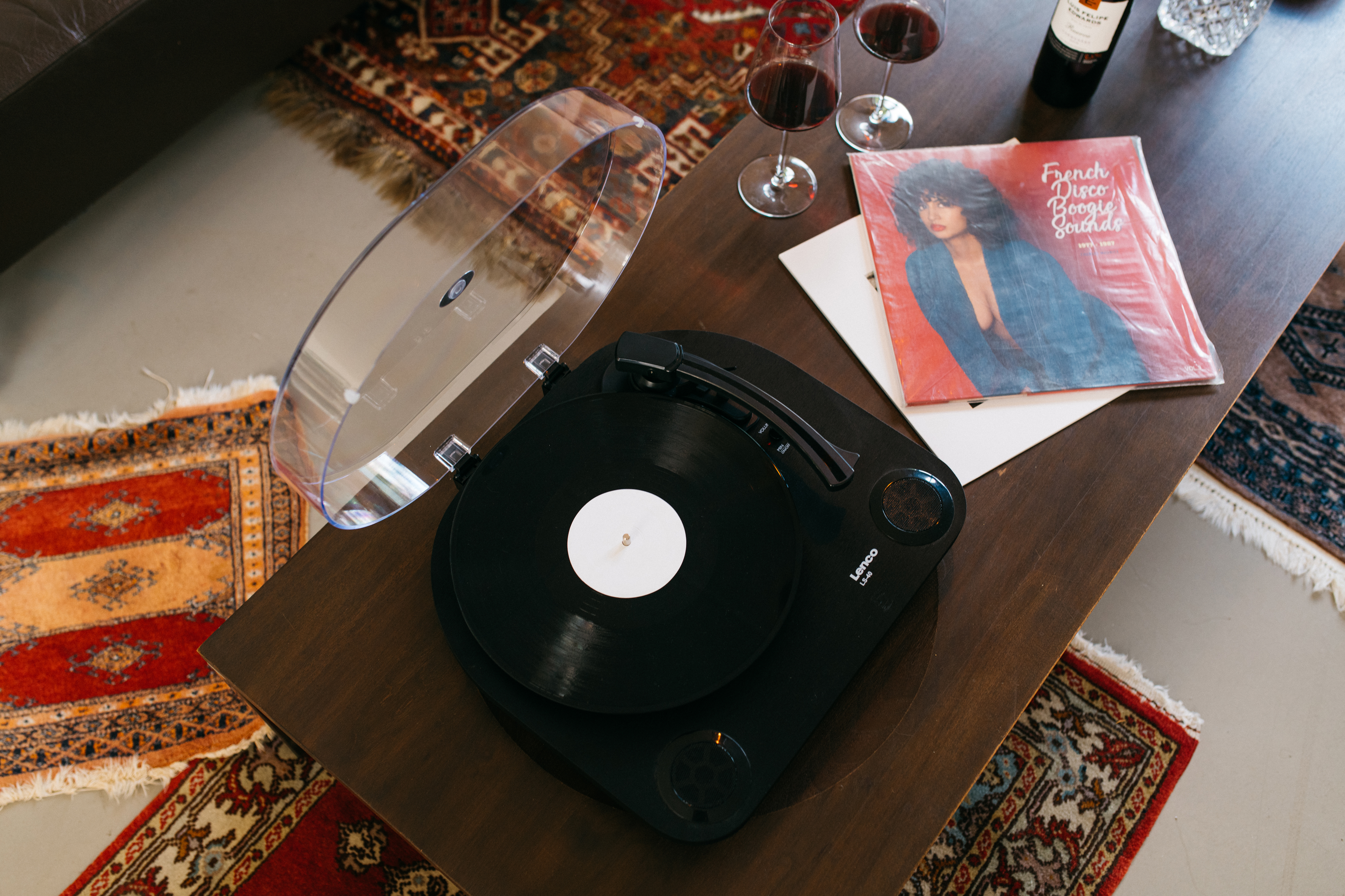 Une platine vinyle sur une table en bois avec des disques, une bouteille de vin et des verres.