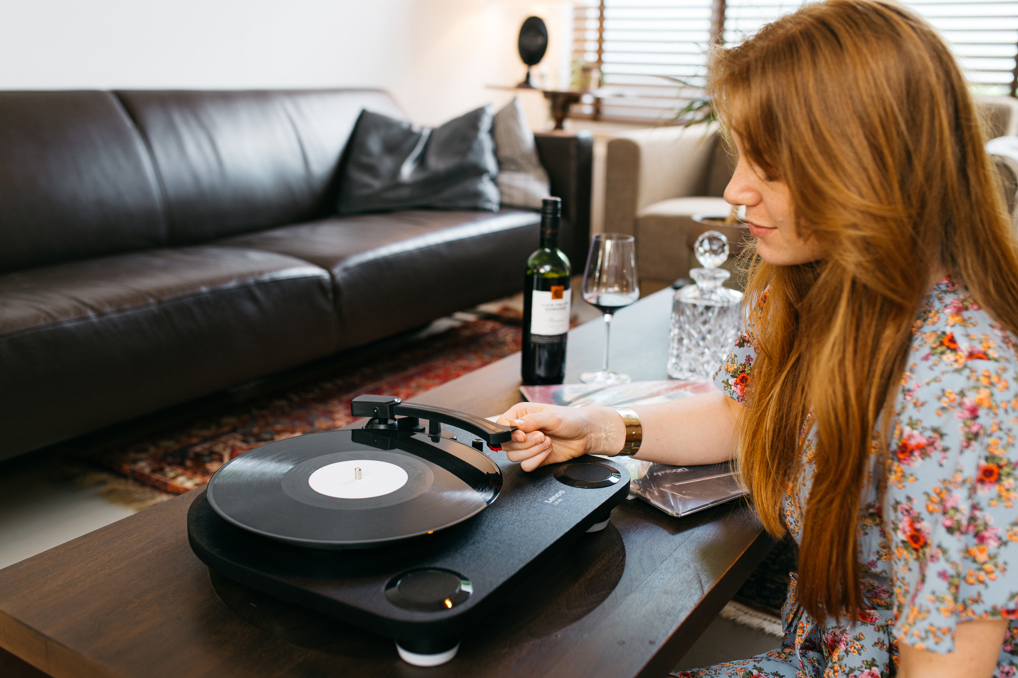 Une femme aux cheveux roux écoute de la musique sur une platine, avec une bouteille de vin et un verre.