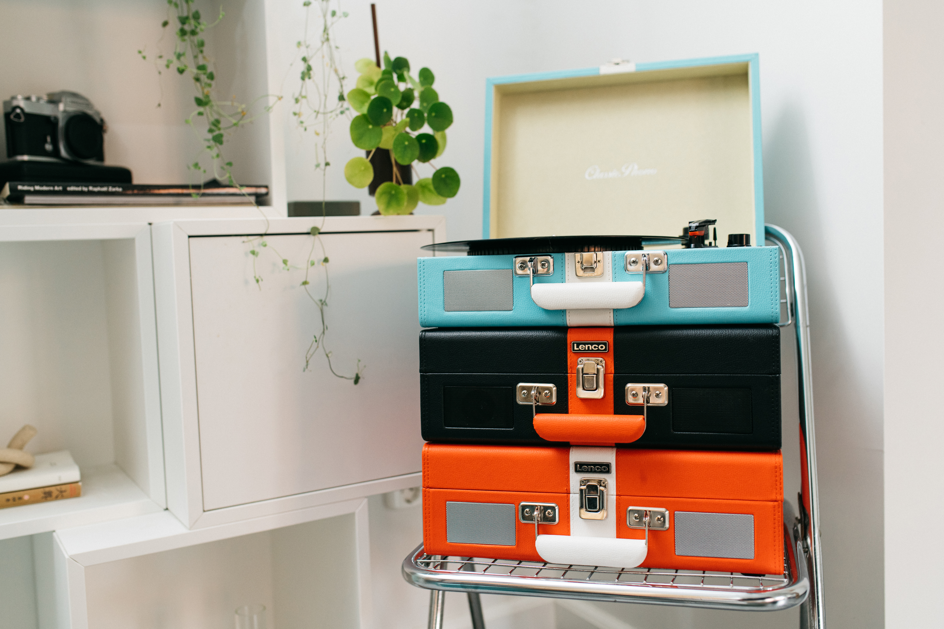 Ułożone w stos gramofony, turkusowy, czarny i pomarańczowy, na metalowym stojaku, z białą półką w tle.