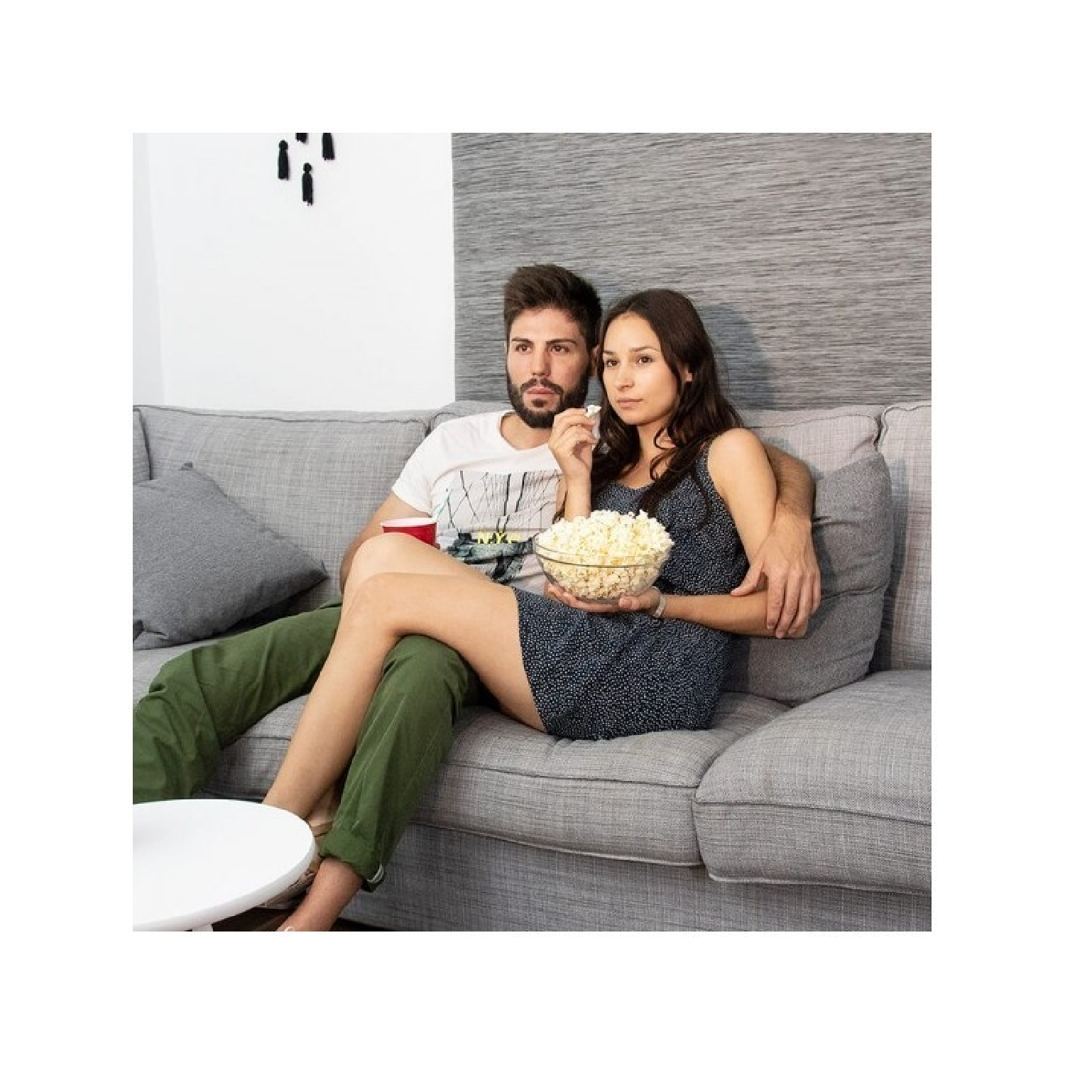 Coppia sul divano, mangiando popcorn. Uomo in maglietta bianca, donna in abito blu. Muro grigio.