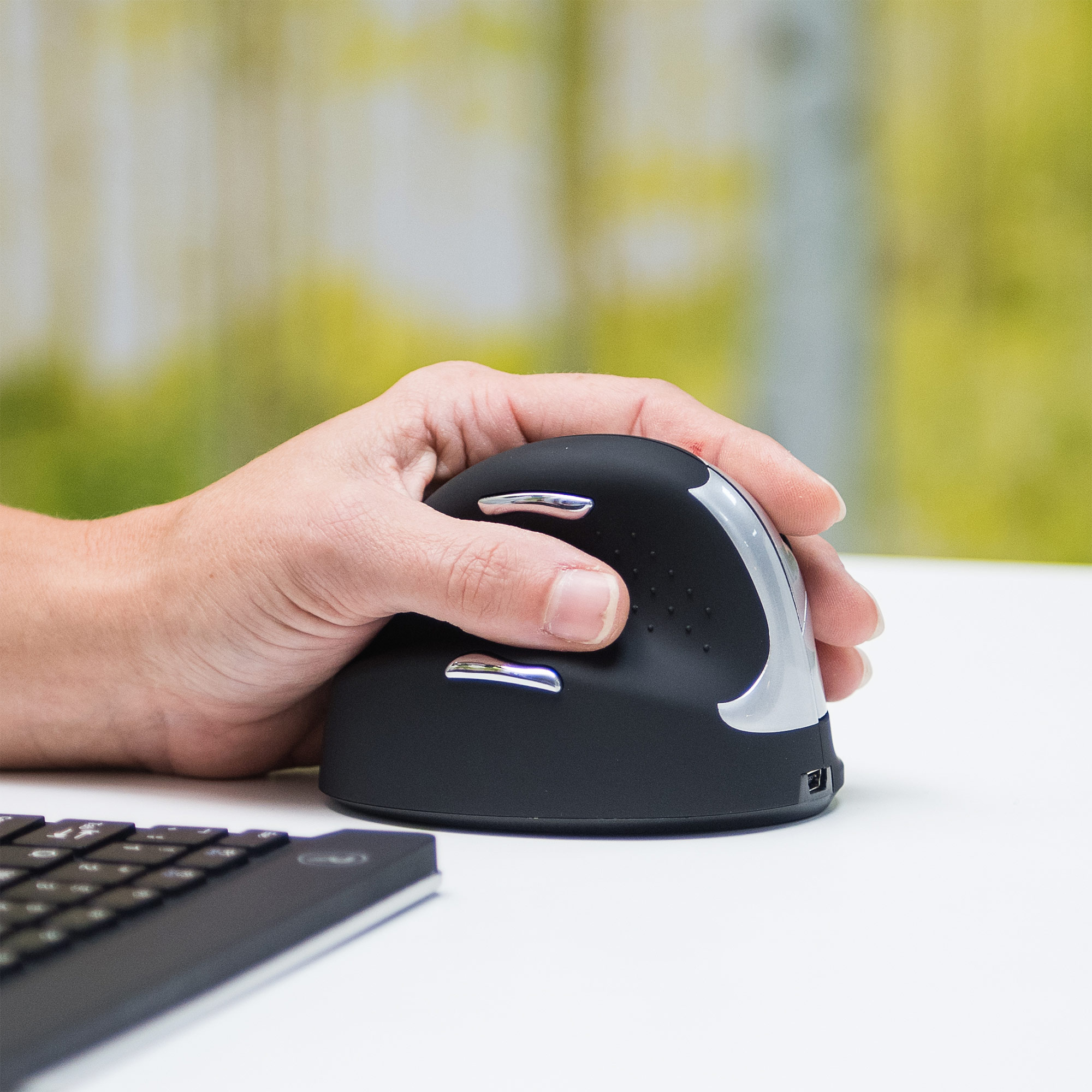 Die Hand einer Person, die eine schwarze, ergonomische, vertikale Maus hält. Eine Tastatur ist teilweise sichtbar.