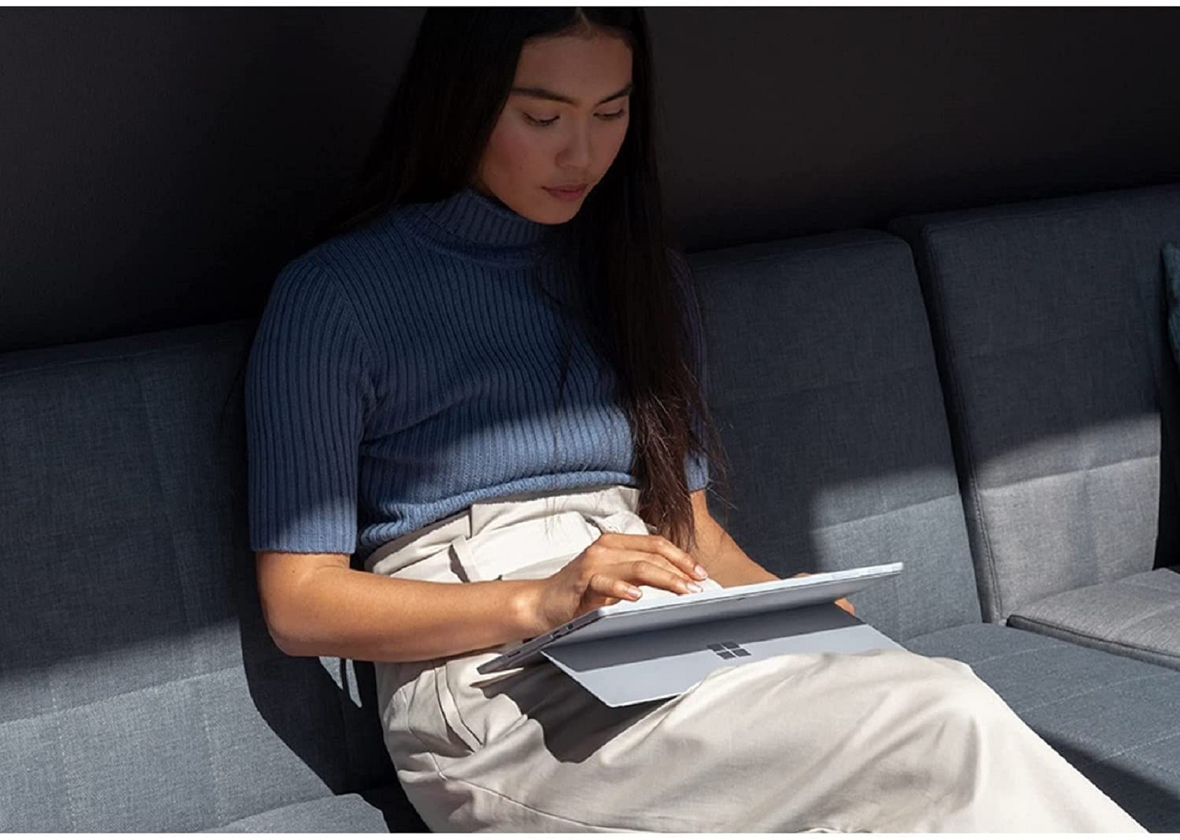 Frau sitzt auf grauer Couch und benutzt Tablet. Sie trägt blaues Oberteil und beige Hose. Natürliches Licht.