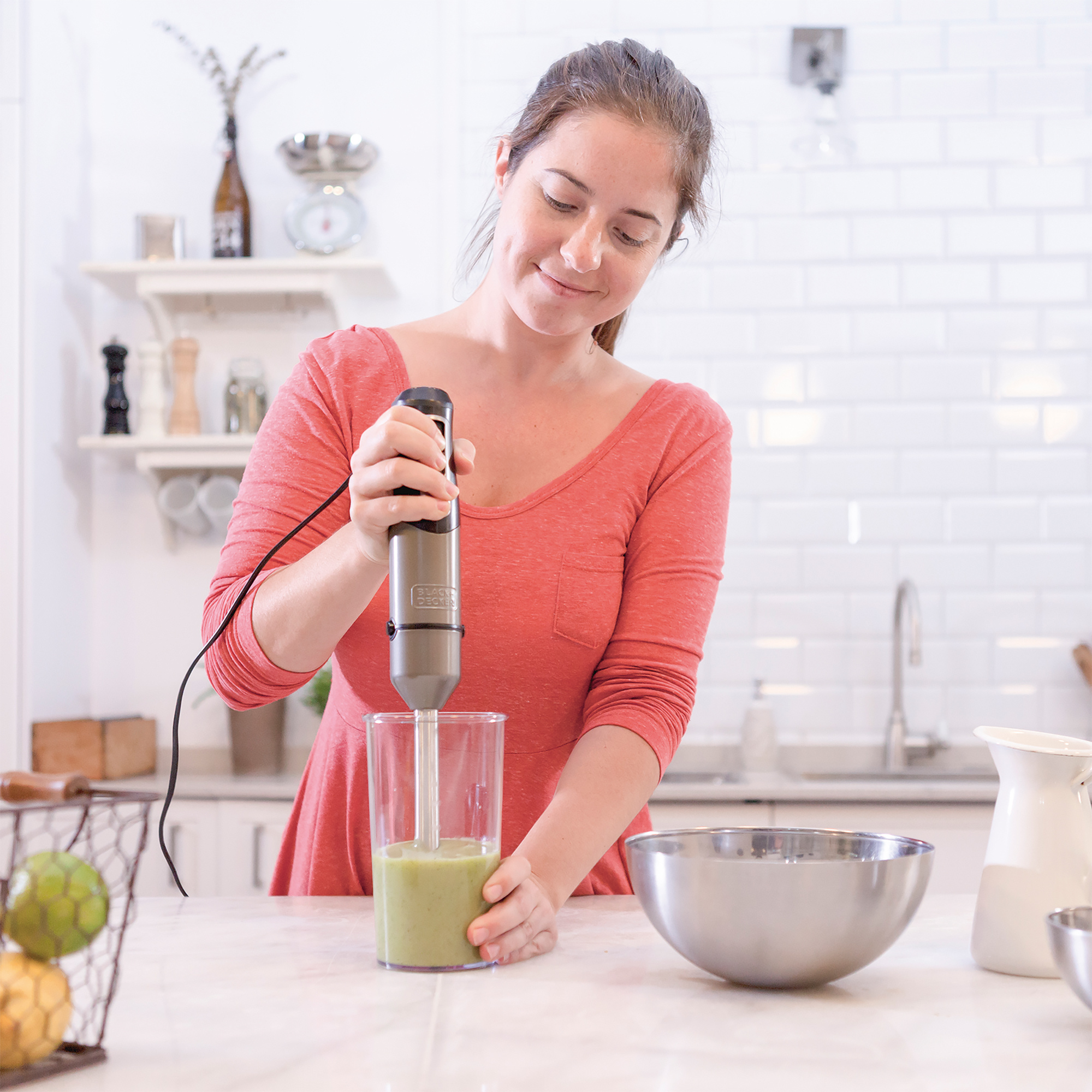Kobieta blenduje zielone smoothie blenderem ręcznym w otoczeniu kuchni.