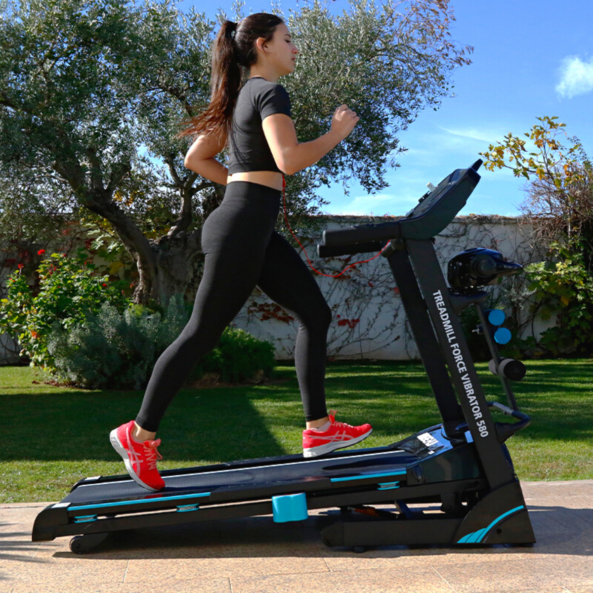 Una mujer corre en una cinta, con ropa deportiva negra y zapatillas rojas, al aire libre.
