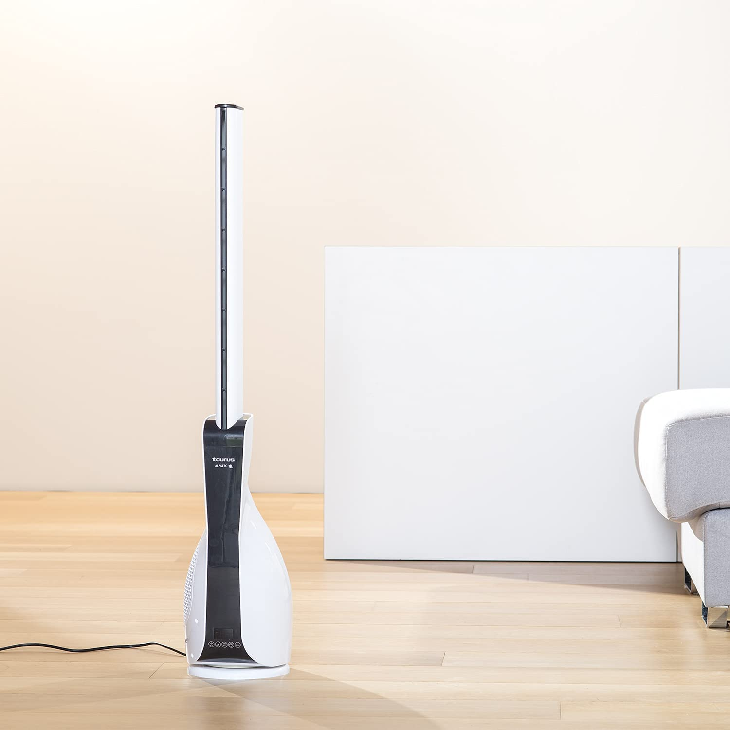 Ventilador blanco y negro. Está sobre un suelo de madera, con una pared blanca y un sofá detrás.