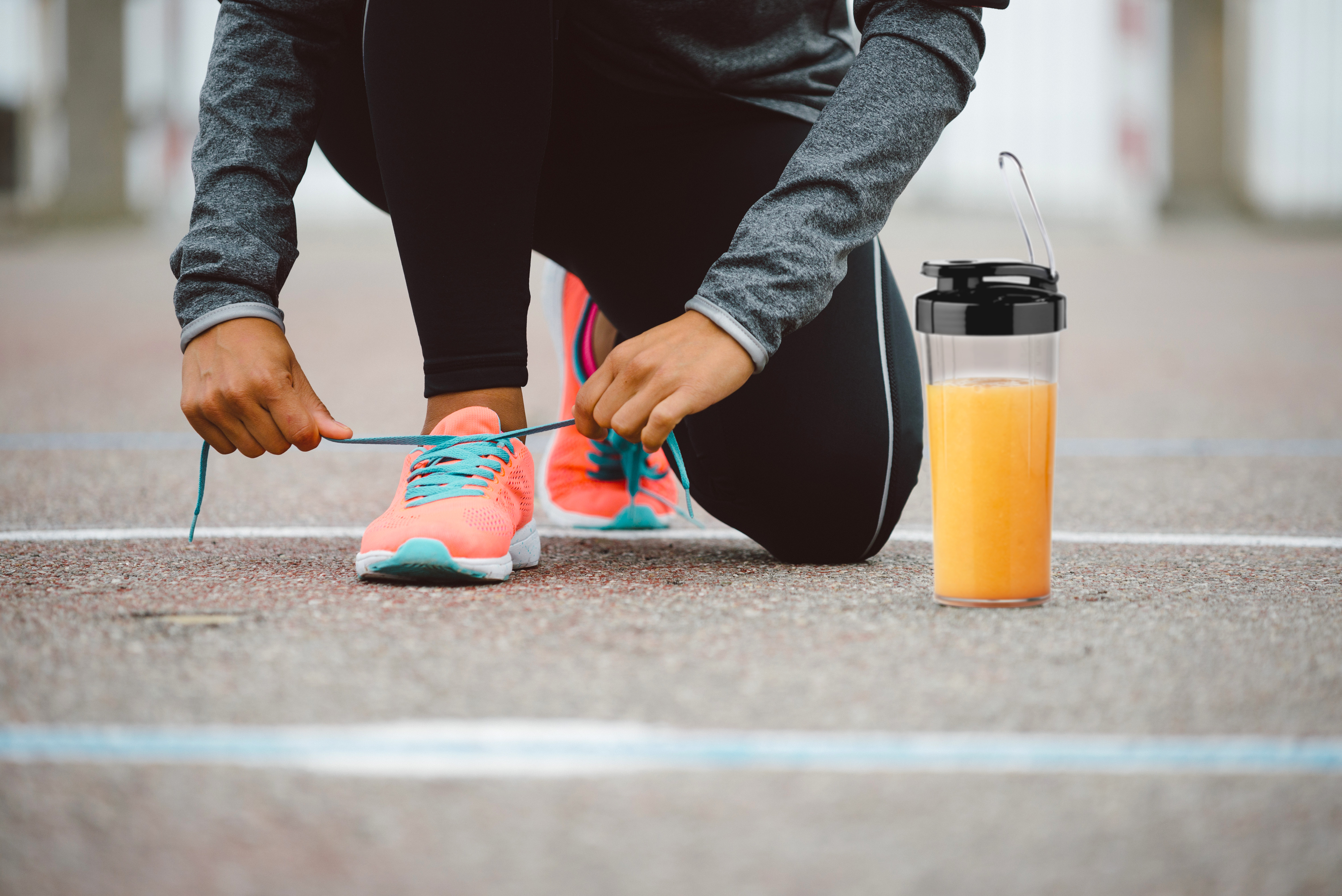 Hardloper strikt veters, oranje en teal schoenen, met een drankje in een shaker. Grijs asfalt.