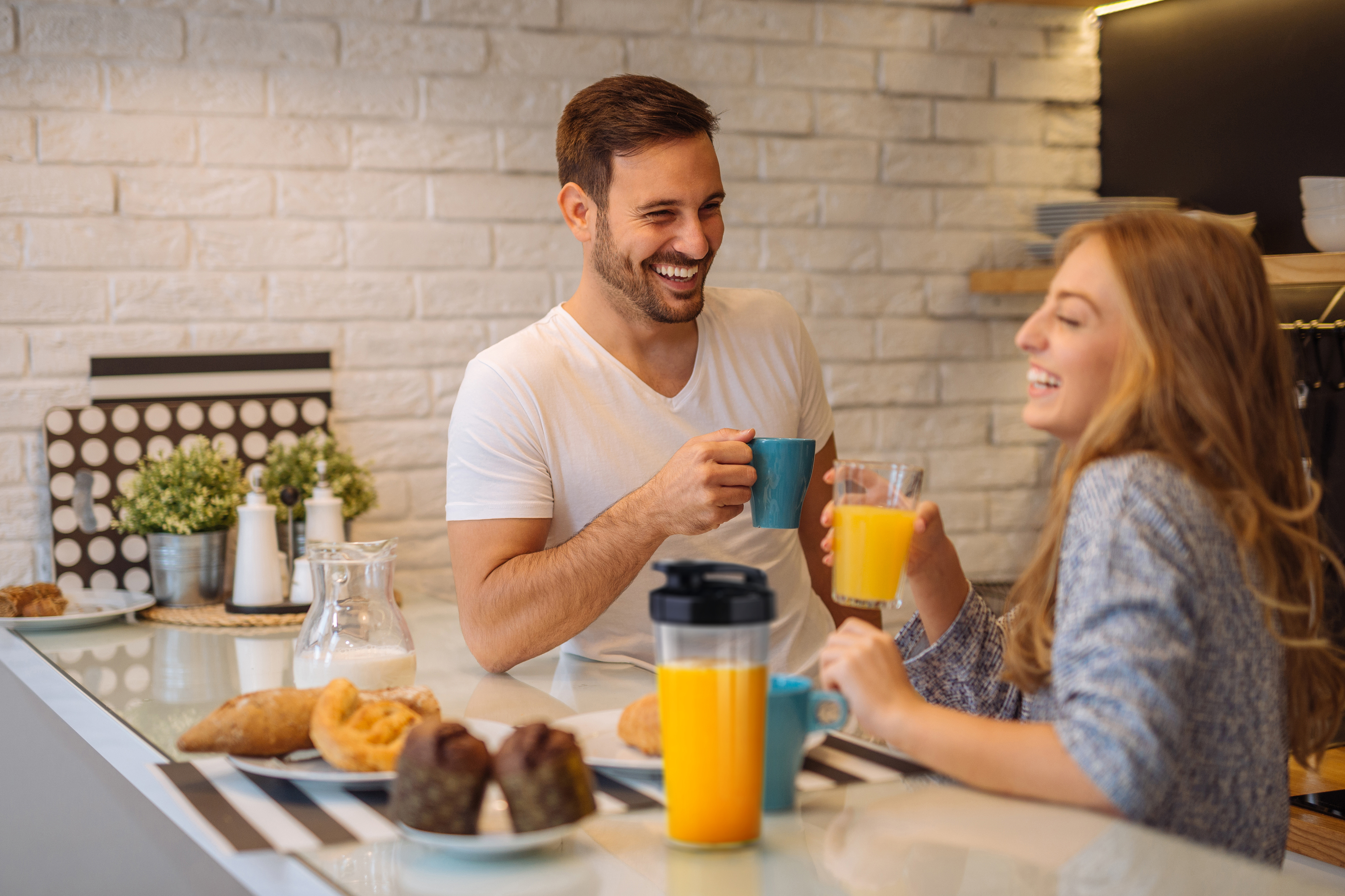 Koppel aan een keukentafel, glimlachend. Man met een mok, vrouw met een sapglas.