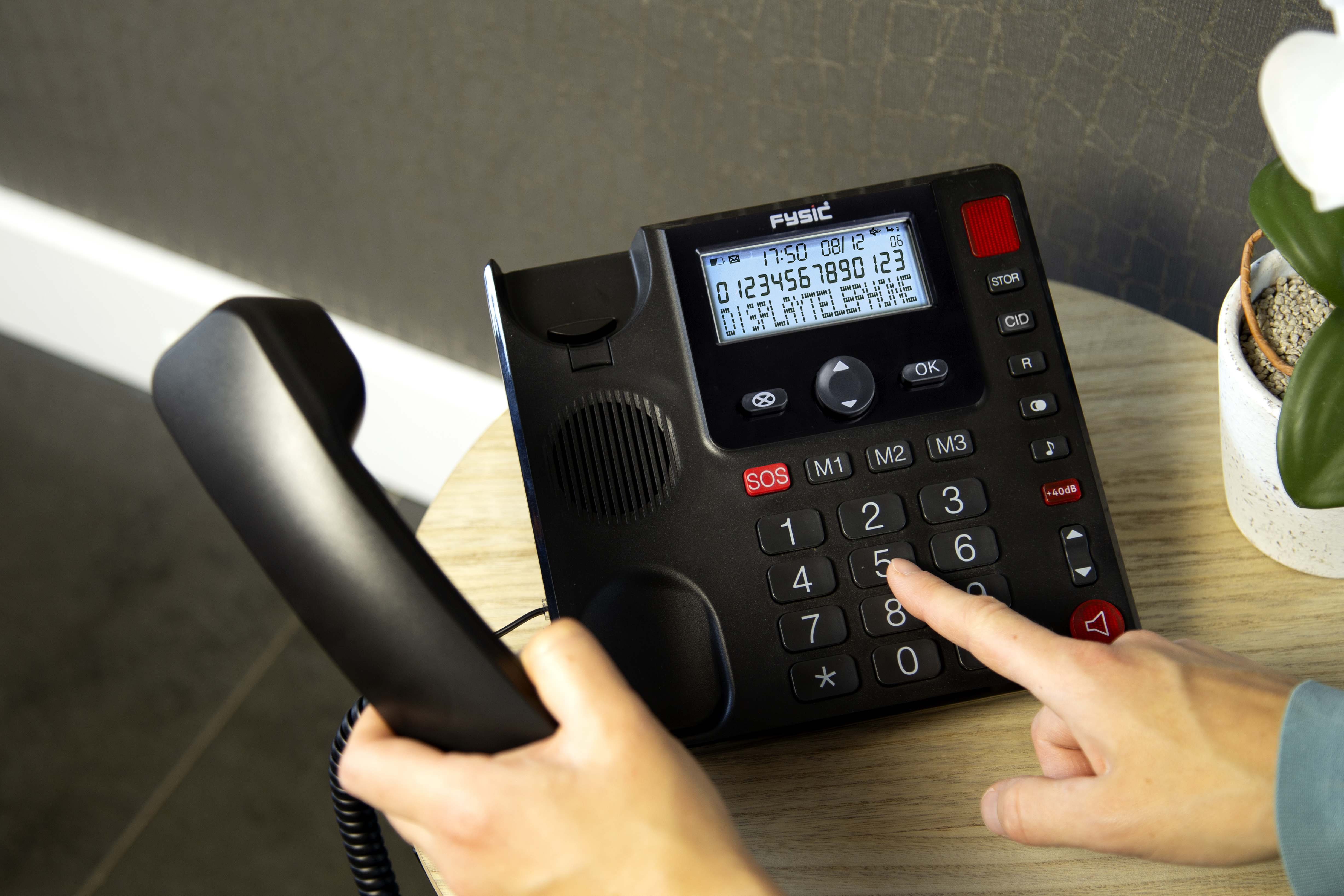 A person's hand dialing a black desk phone with a display showing numbers, on a wooden table.