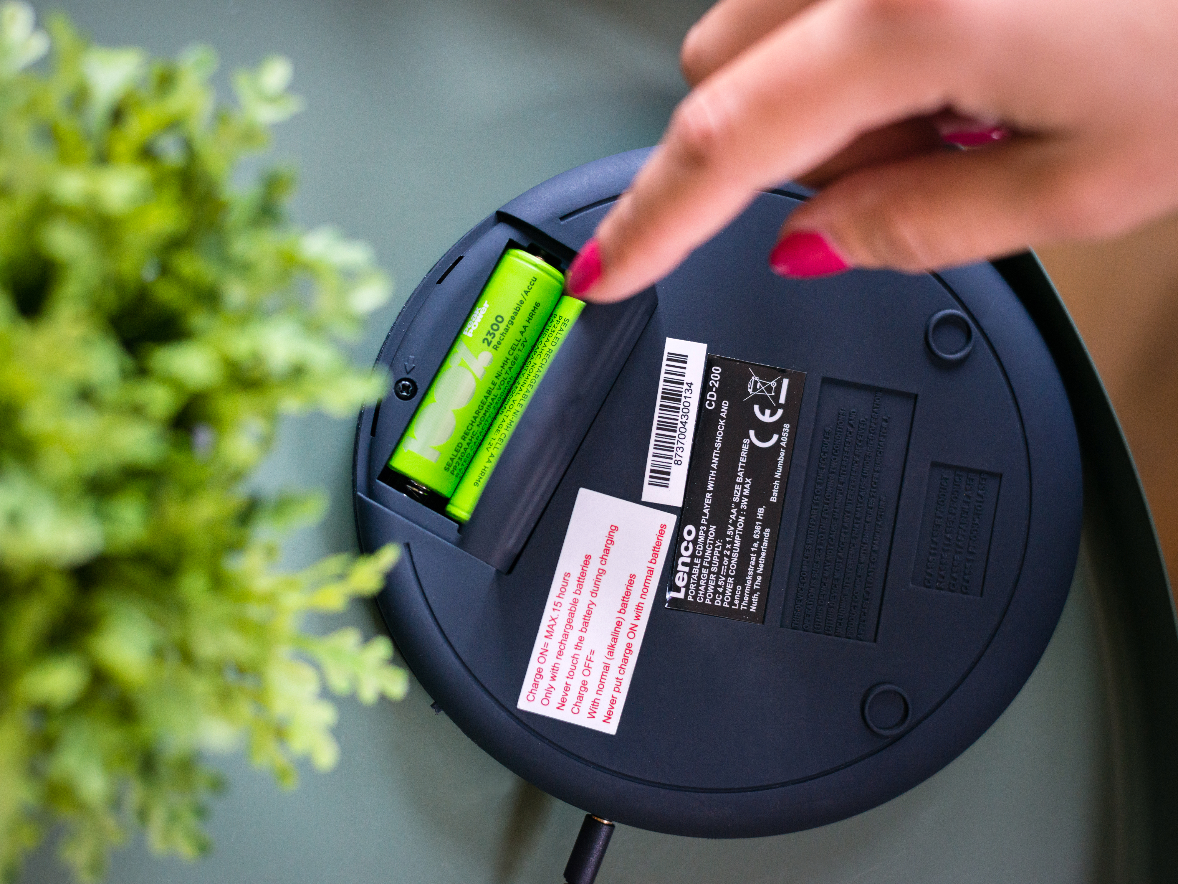 Osoba wkłada zieloną baterię do czarnego odtwarzacza CD. Tło to zielony stół i rośliny.