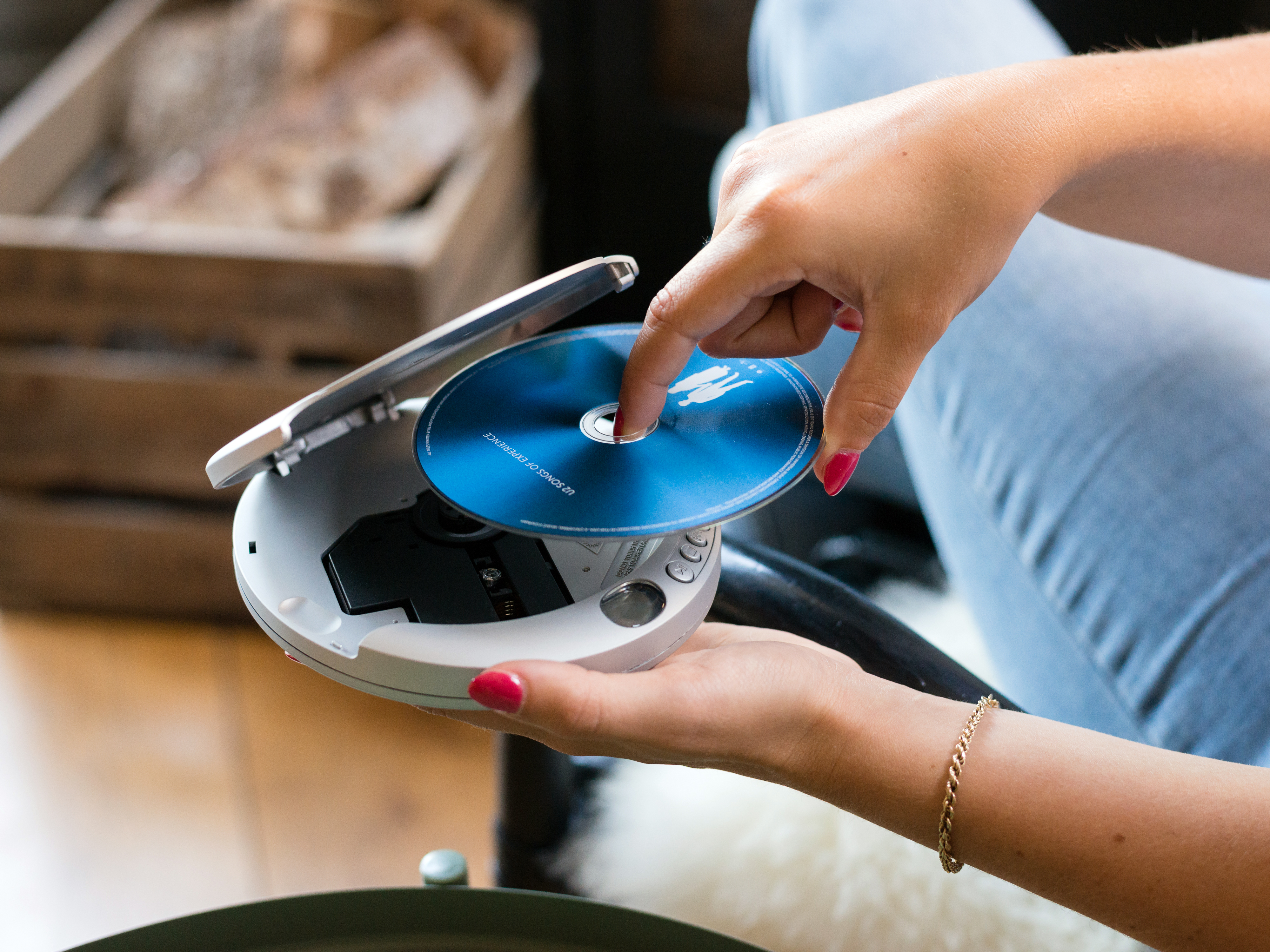 Osoba wkłada płytę CD do białego, otwartego odtwarzacza CD. Z tyłu drewniana skrzynia i dywan.