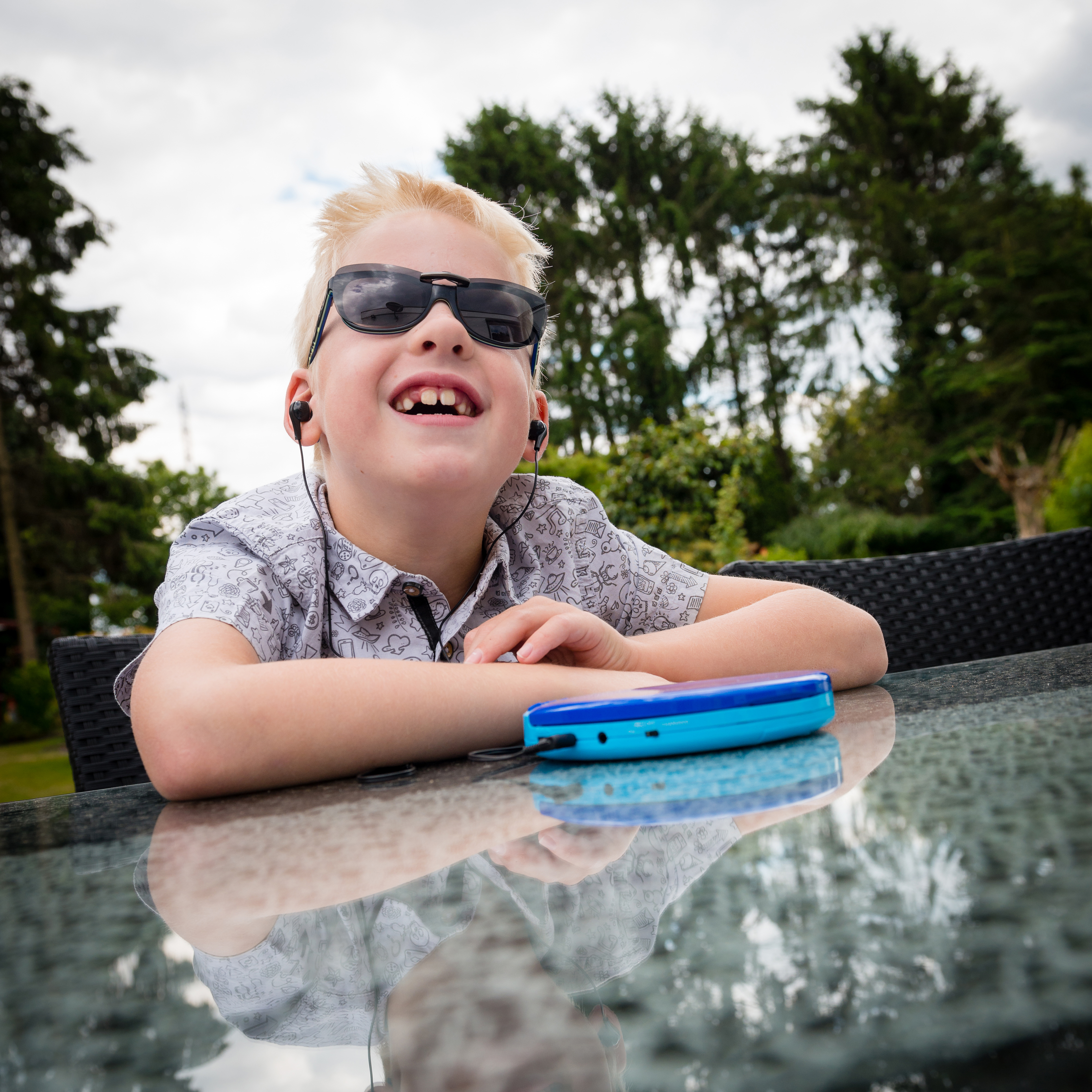 Chłopiec w okularach przeciwsłonecznych i słuchawkach uśmiecha się patrząc w górę. Na stole niebieski odtwarzacz muzyki.