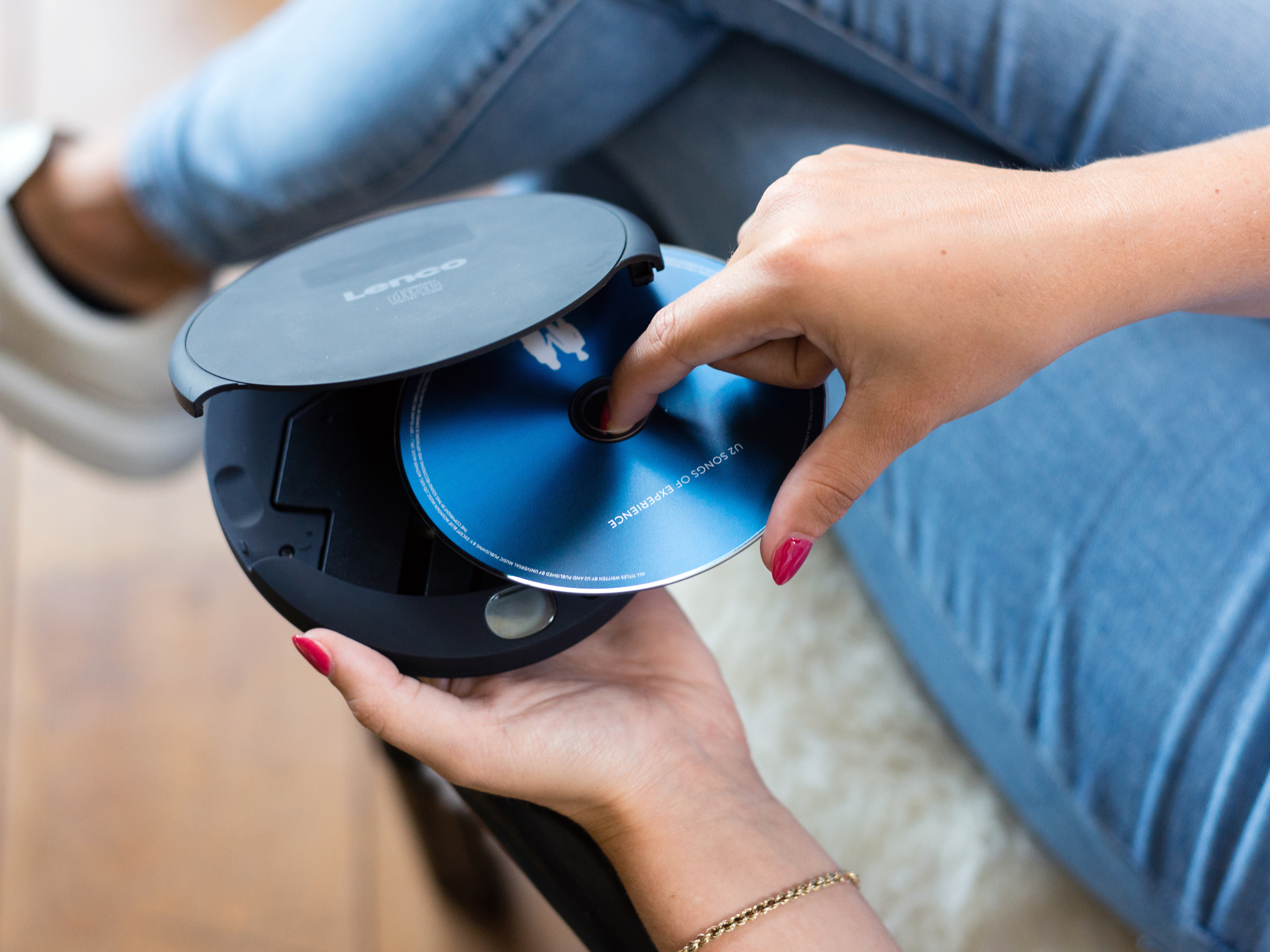 Osoba wkłada płytę CD do czarnego odtwarzacza CD Lenco. W tle widoczne są dżinsy i beżowe buty.