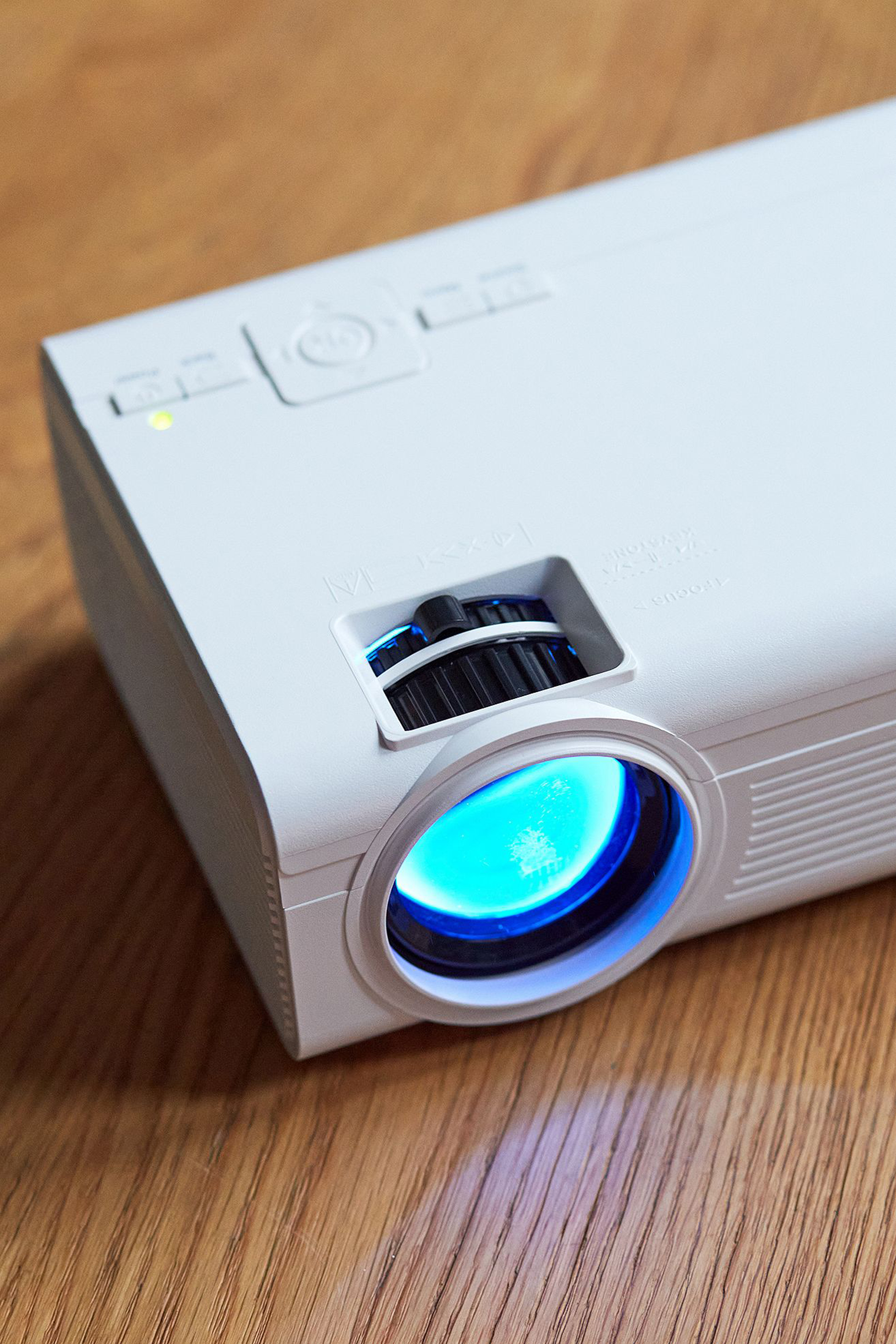 Biały projektor z niebieskim światłem z obiektywu, umieszczony na drewnianej powierzchni.