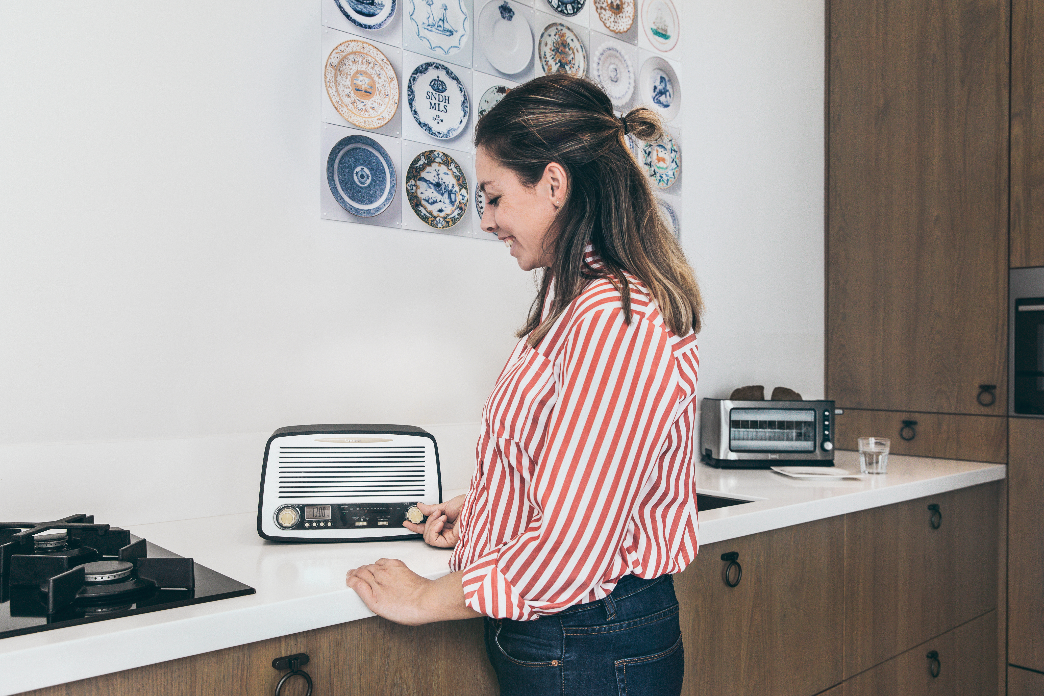 Woman in a kitchen, turning a radio dial. A toaster and plates on the wall in the background.