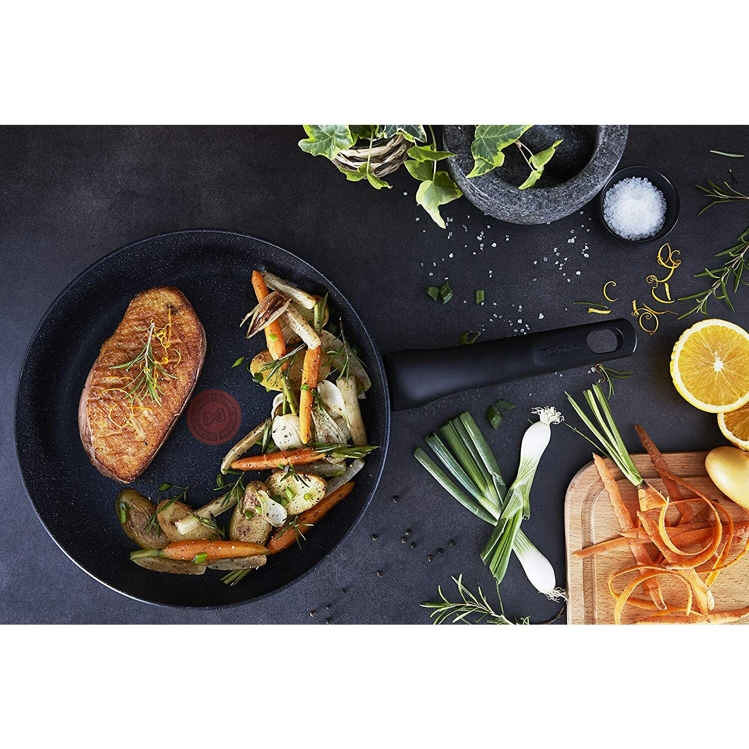 Patelnia z jedzeniem: stek, warzywa, zioła i plasterki pomarańczy. Czarne tło.